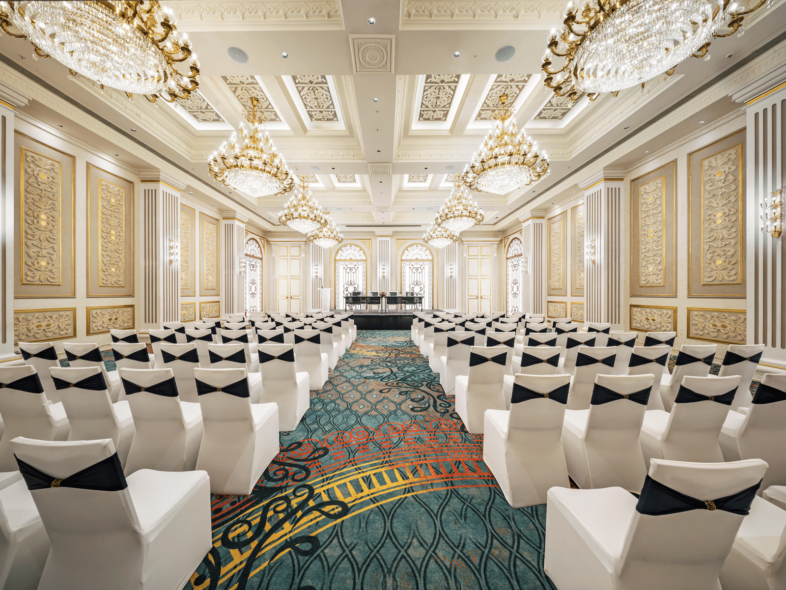 A grand hall prepared for a reception, showcasing elegant round tables with golden accents and white chairs, all beneath a ceiling adorned with multiple sparkling chandeliers.