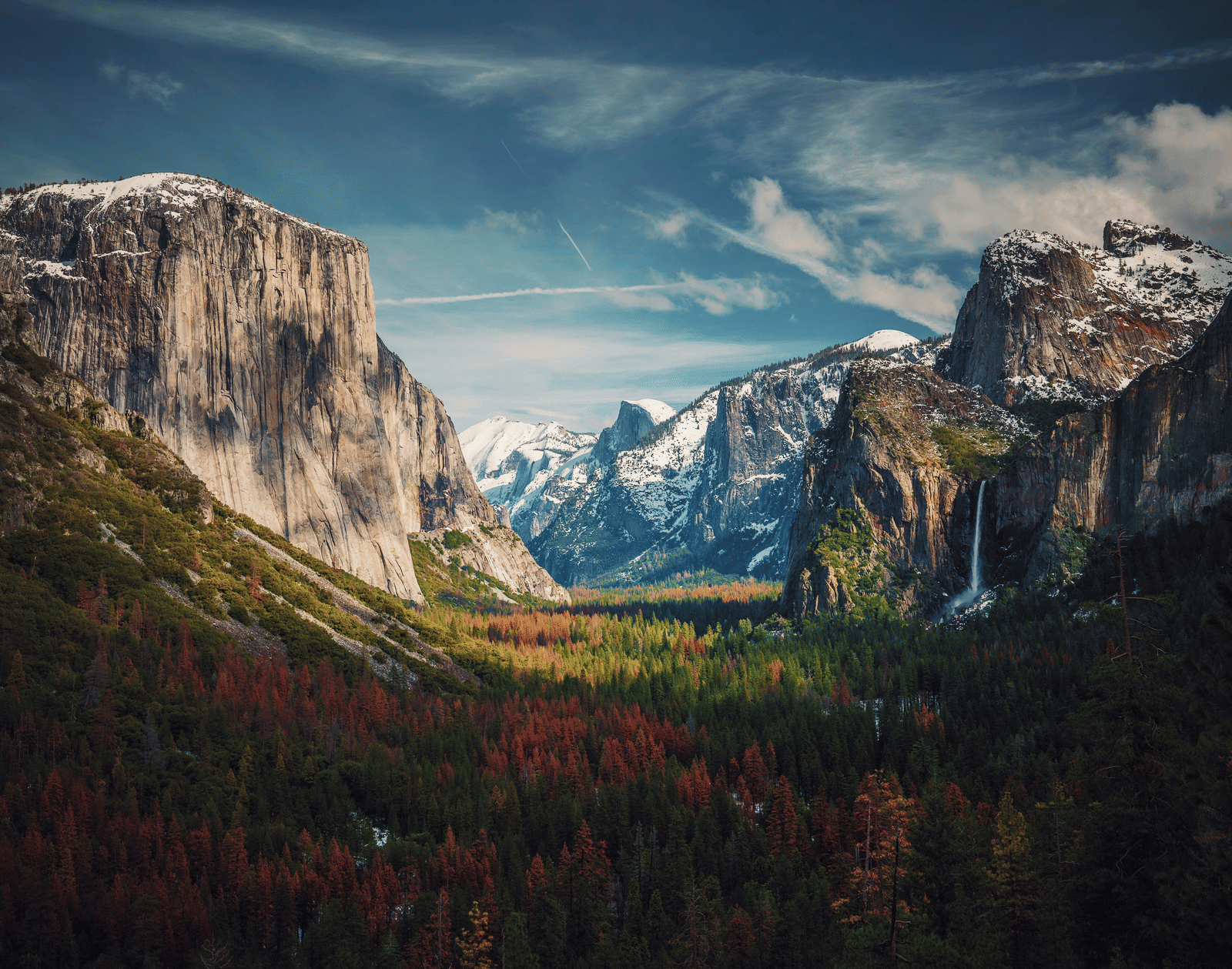 A dramatic view of Yosemite Valley with towering granite cliffs, including El Capitan, snow on the peaks, and autumn colors in the forest.