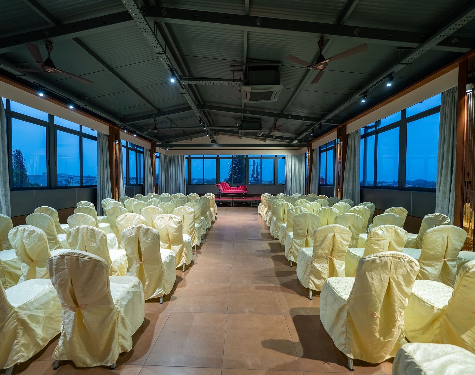 A view of the banquet hall at Hari Govinda's Express, Vijayanagar, with chairs arranged on either side of a central aisle leading to the stage, which features a cushioned sofa, while the curtains are drawn to the sides.