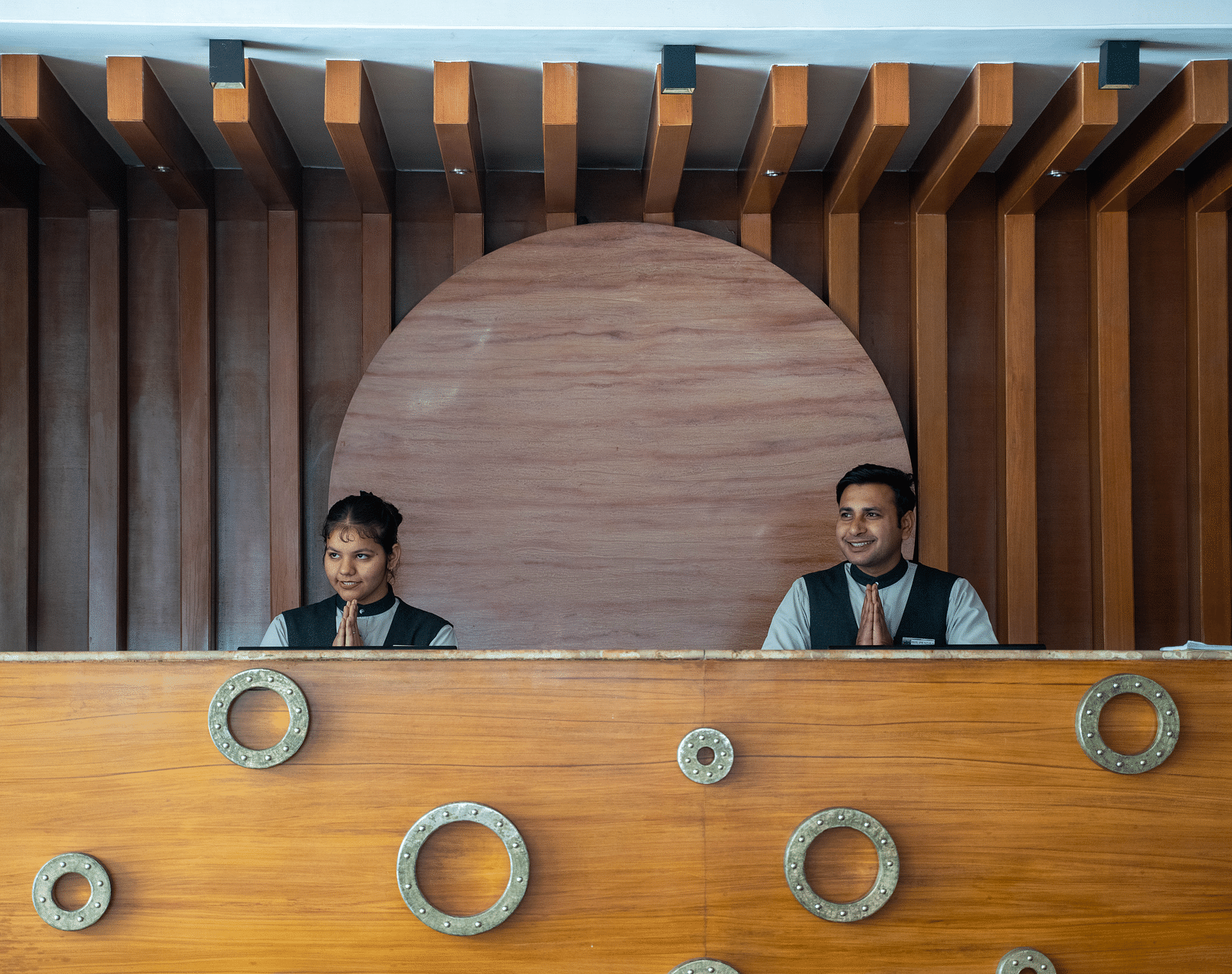 Two hotel staff members standing behind a stylish wooden reception counter with circular design accents - Hotel Udai Median, Udaipur.
