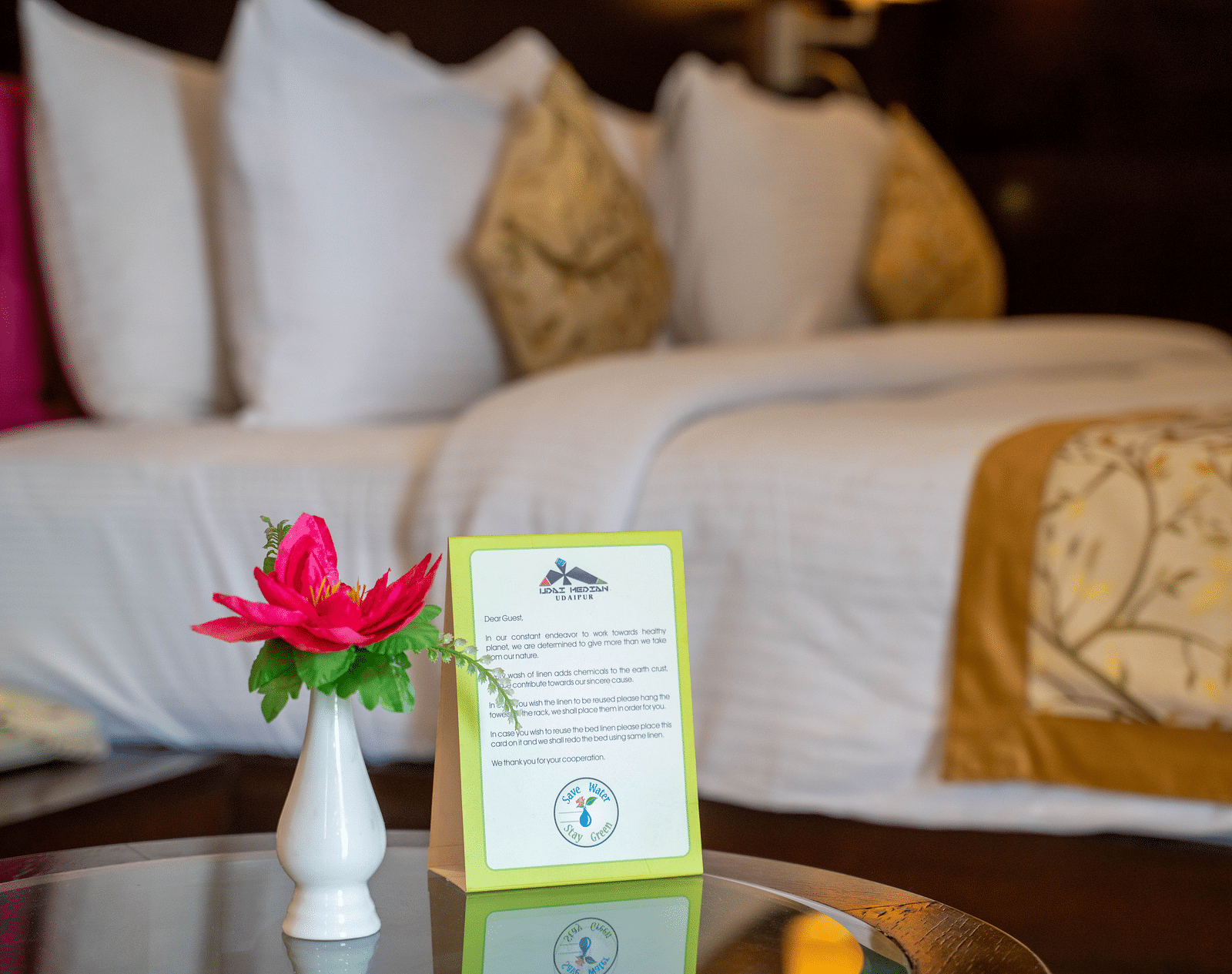 A neatly made hotel bed with white linens, decorative pillows, and a bedside table holding a flower vase and a service card - Hotel Udai Median, Udaipur.
