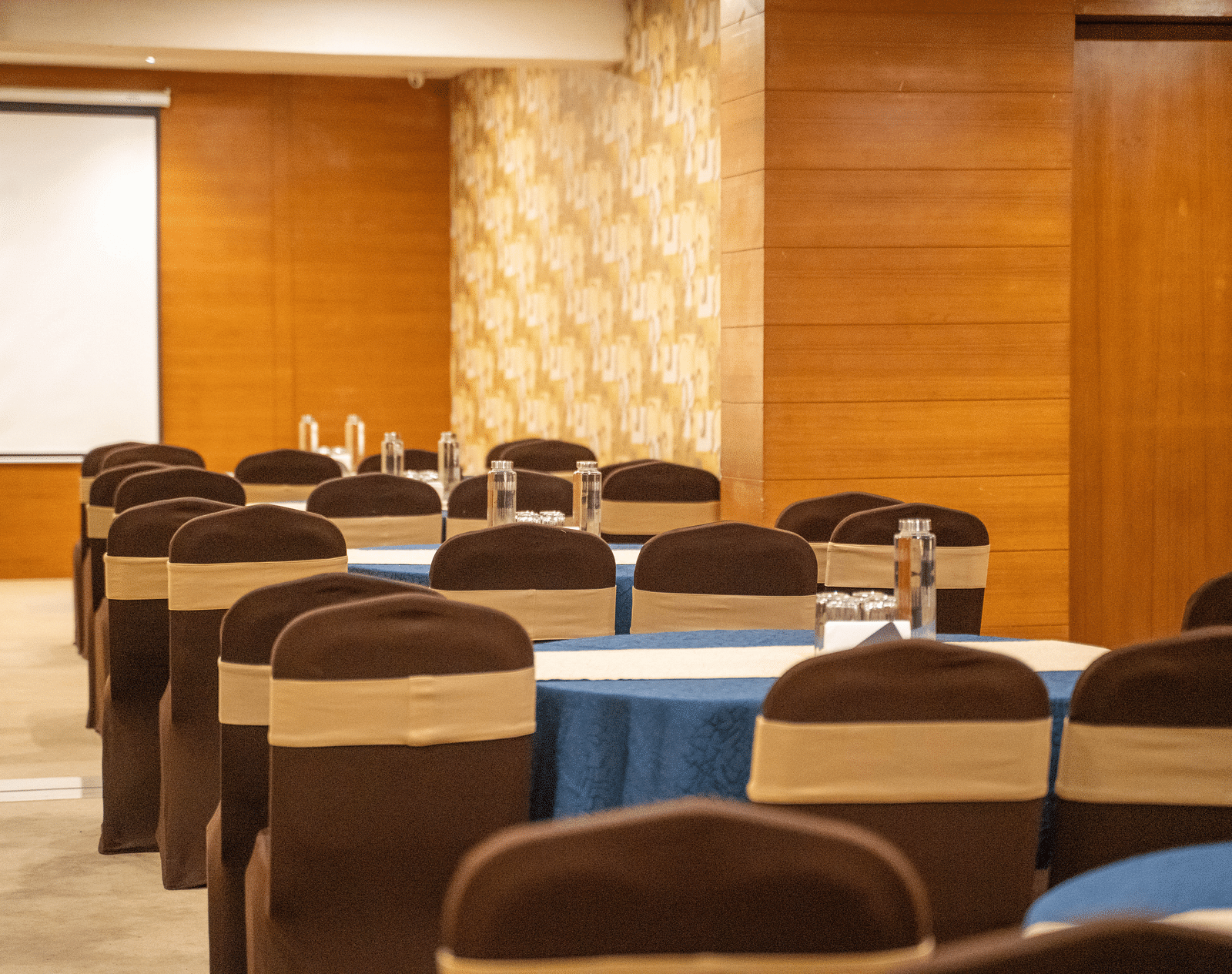 A spacious conference room with rows of round tables, upholstered chairs, and a presentation screen - Hotel Udai Median, Udaipur.