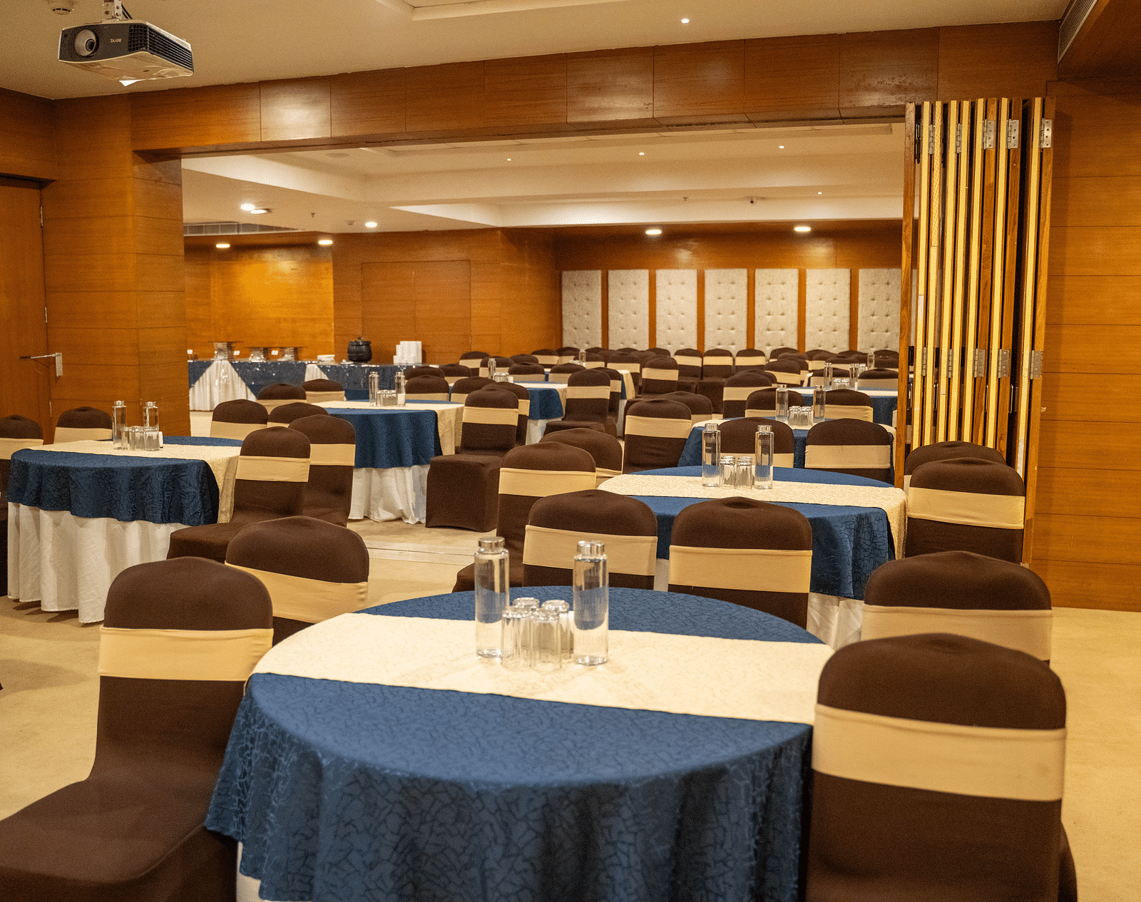 A banquet hall with round tables draped in blue tablecloths and brown chairs, arranged for an event - Hotel Udai Median, Udaipur.