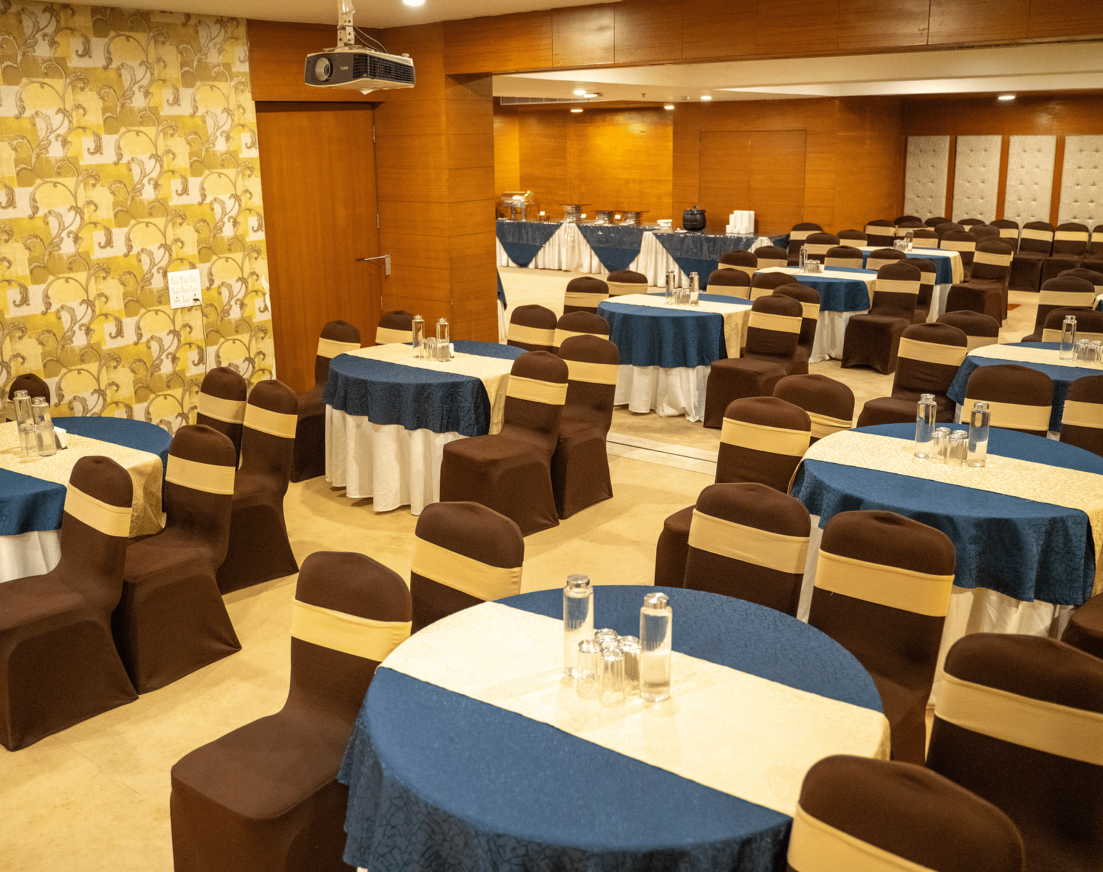 A well-lit event space with round tables, a wooden backdrop, and warm decor - Hotel Udai Median, Udaipur.