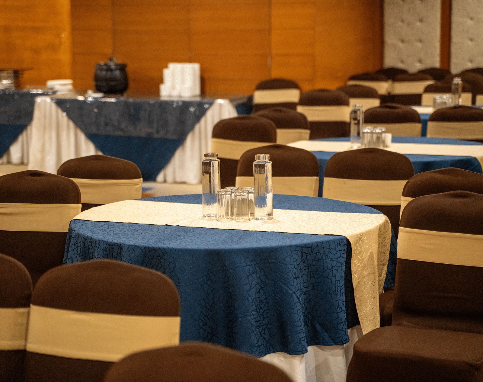 A banquet hall set up for an event, with neatly arranged round tables covered in blue tablecloths and brown chairs - Hotel Udai Median, Udaipur.