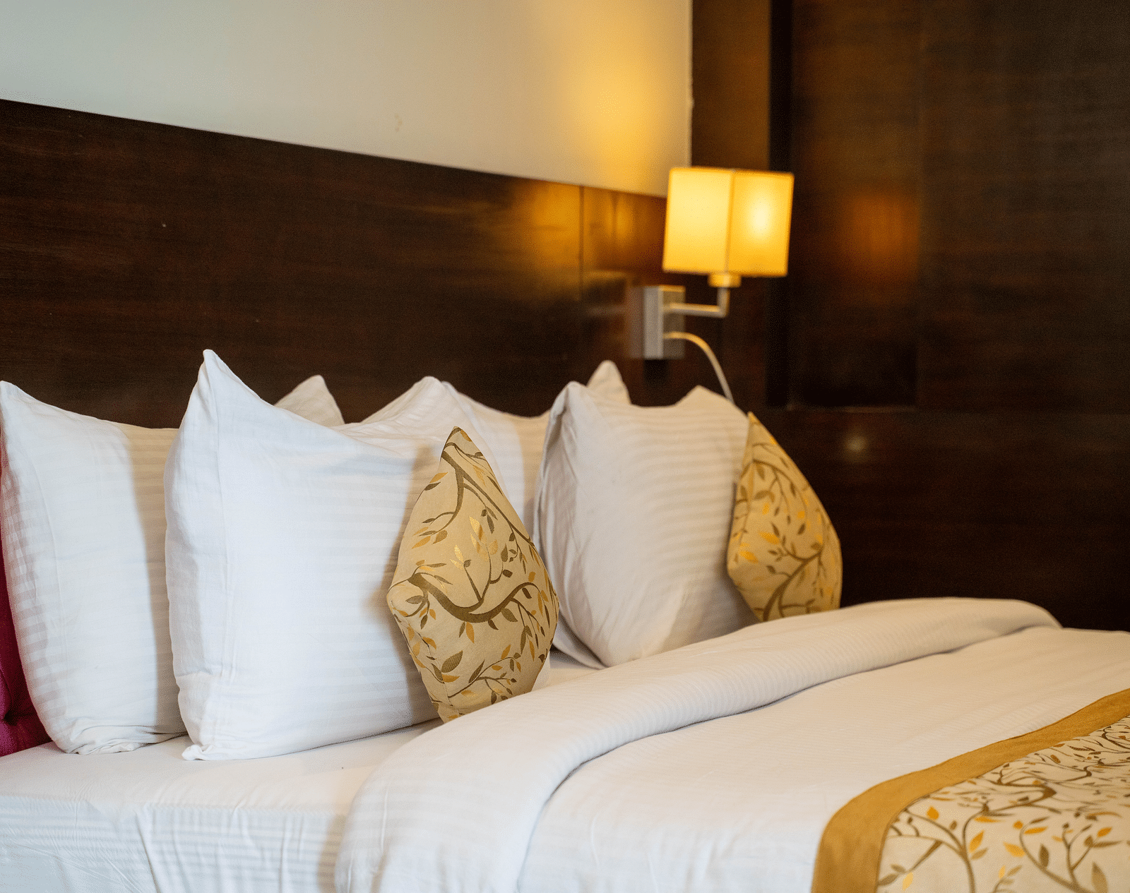 A neatly made bed with white linens and multiple pillows, including a patterned decorative pillow, is situated against a dark wood headboard, with a wall-mounted lamp emitting a warm light | Hotel Udai Median, Udaipur