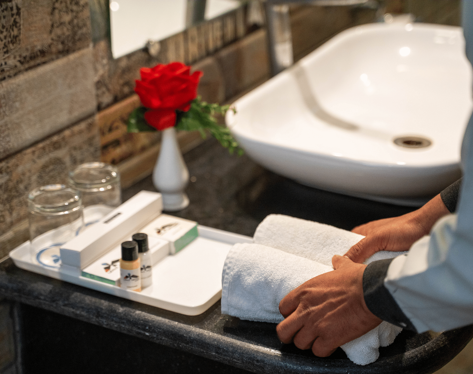 A bathroom countertop shows a white sink and a small vase with a red flower, alongside a tray holding toiletries and two glasses | Hotel Udai Median, Udaipur