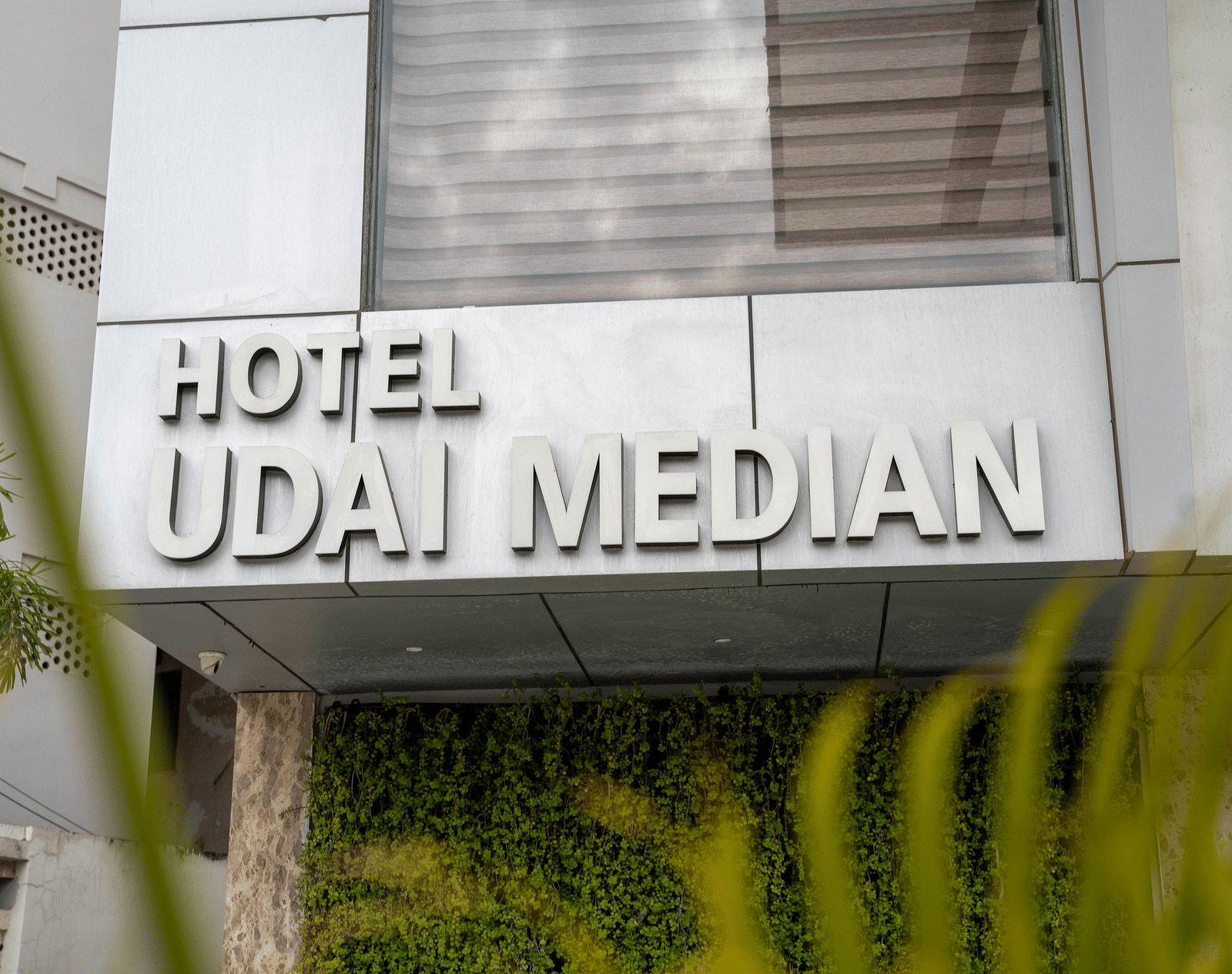 Facade of Hotel Udai Median with a textured wall and greenery visible below.