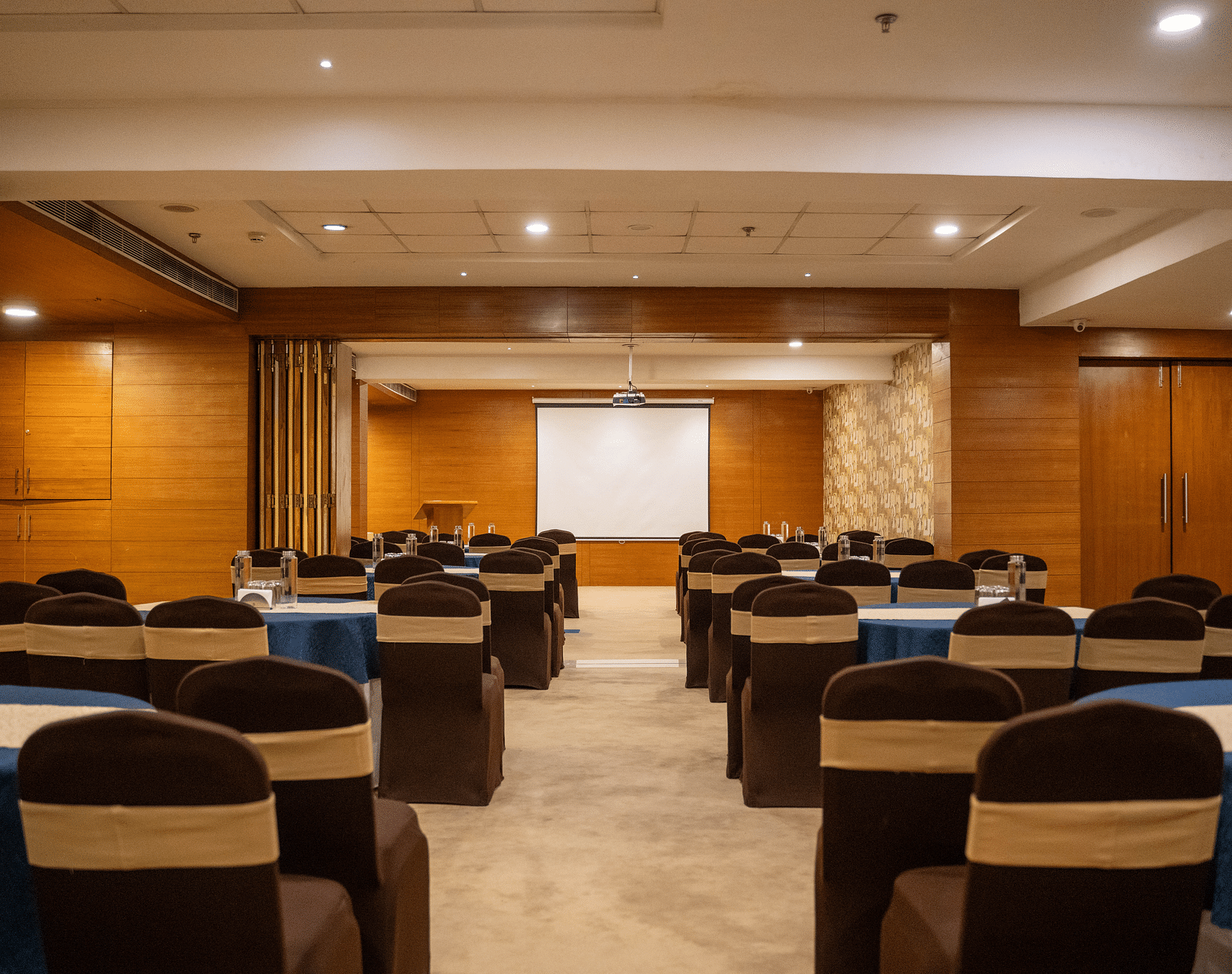 A well-lit, empty conference room features rows of dark brown chairs with light tan sashes, arranged around blue-clothed tables, facing a blank white projector screen at the room's far end | Hotel Udai Median, Udaipur