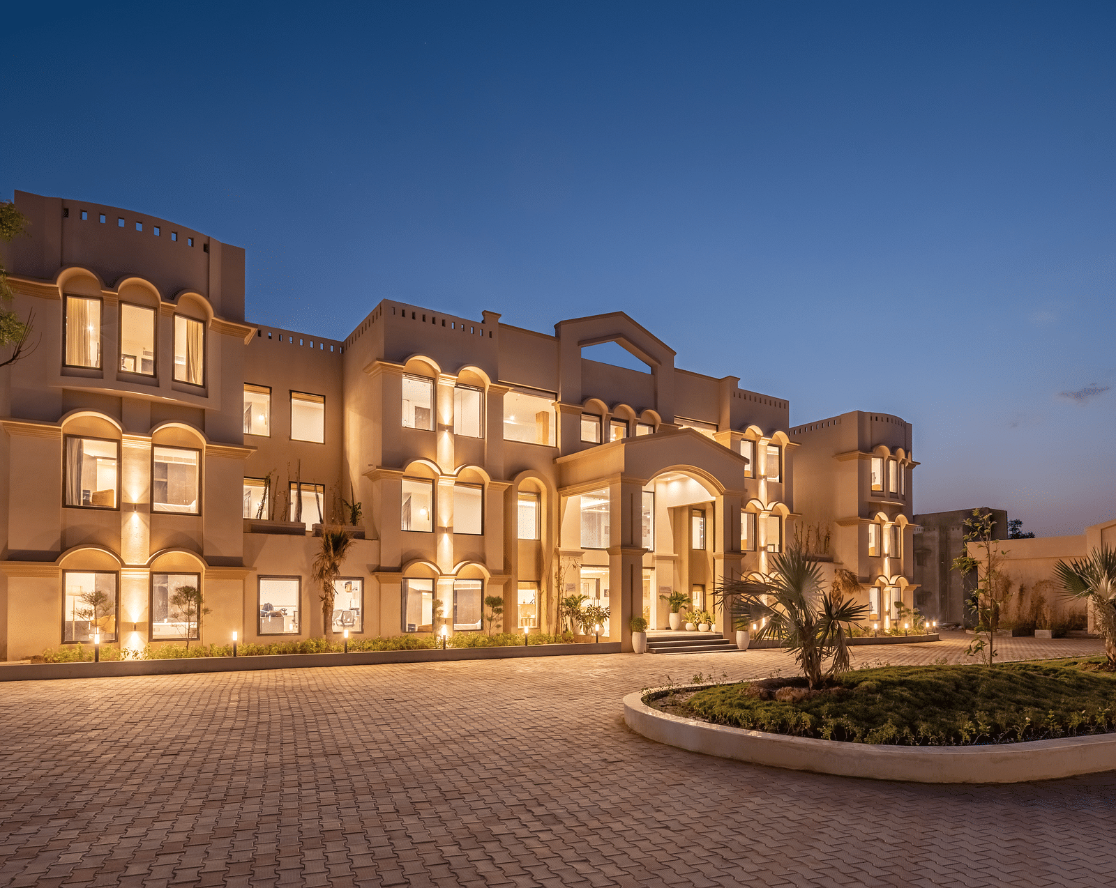 An image of a well lit facade with a huge concrete lawn and a few trees - Brij Eternity, Vrindavan