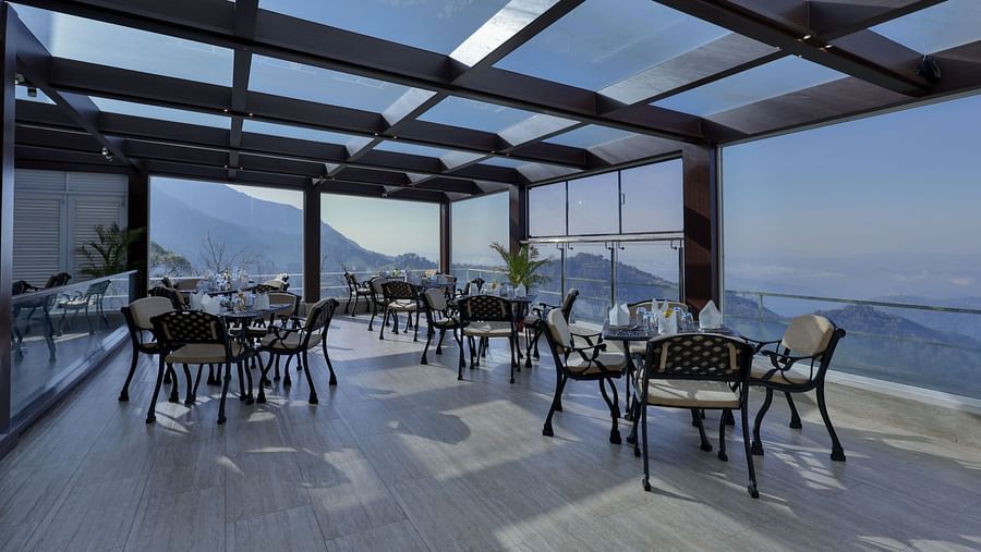 Scenic interior of a restaurant with tables and chairs, offering panoramic glass views of the mountains – Fragrant Nature Munnar.