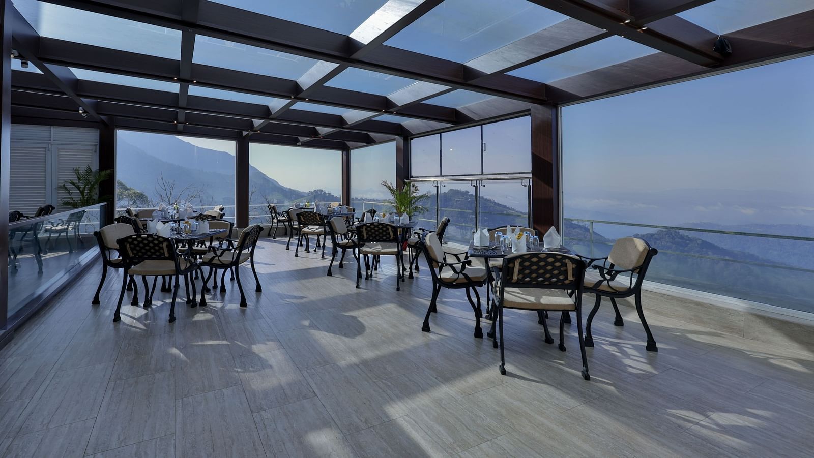 Scenic interior of a restaurant with tables and chairs, offering panoramic glass views of the mountains – Fragrant Nature Munnar.