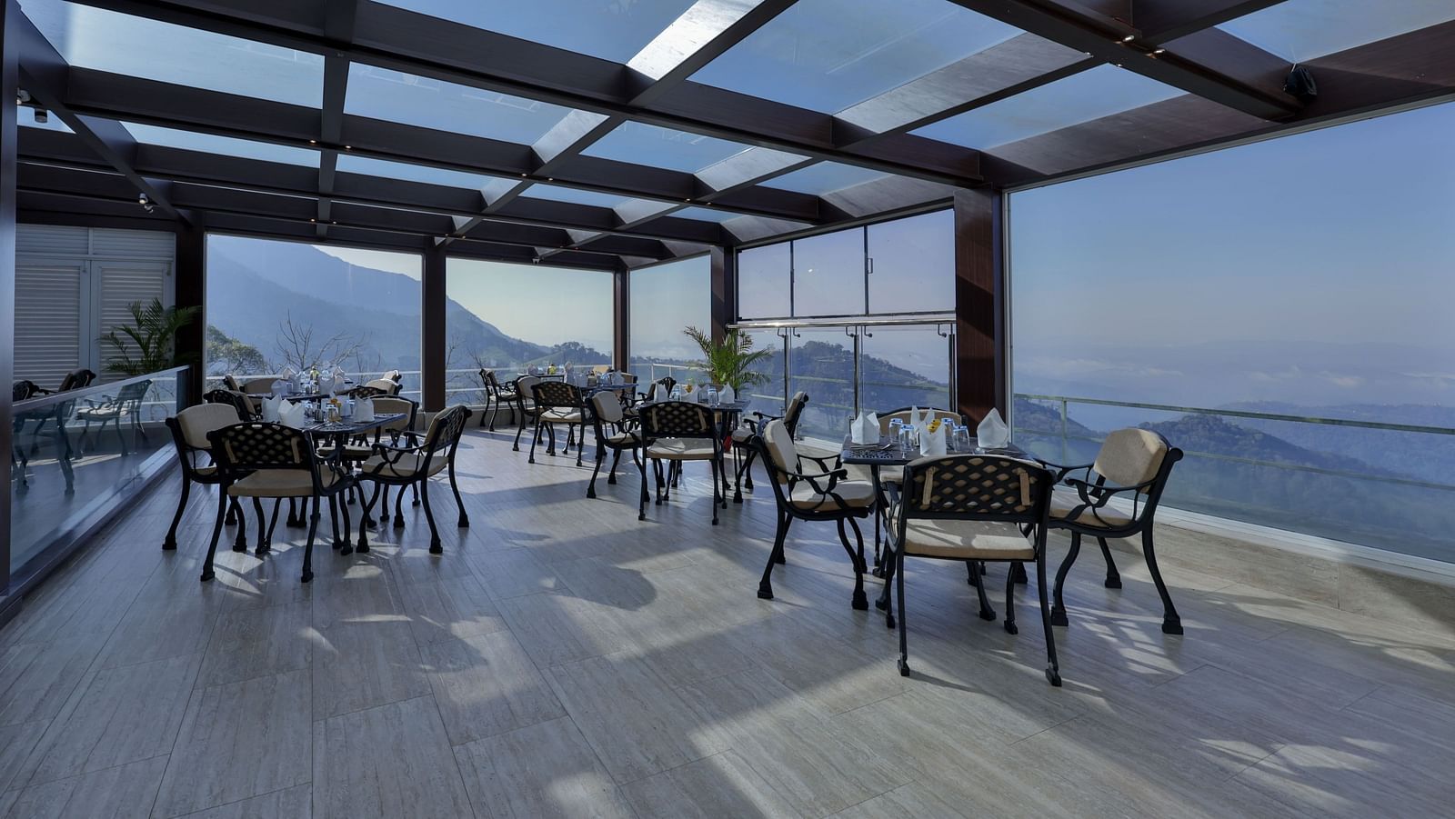 Scenic interior of a restaurant with tables and chairs, offering panoramic glass views of the mountains – Fragrant Nature Munnar.