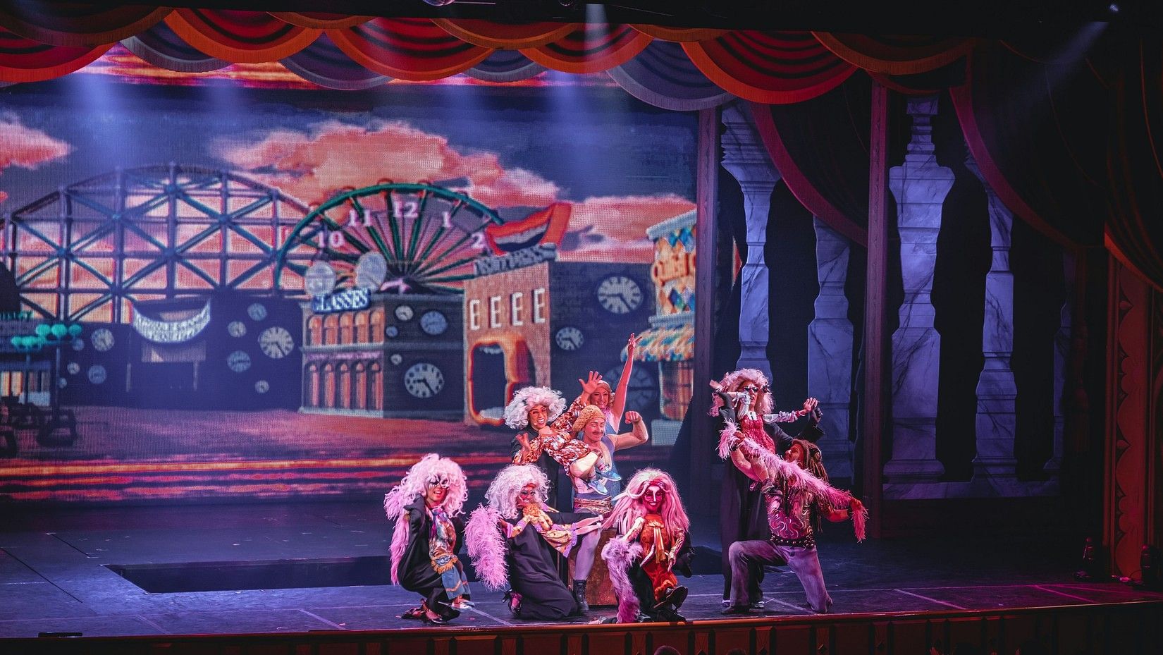 Performers in colorful costumes and wigs act on a vibrant theater stage with a lively amusement park backdrop and dramatic lighting.