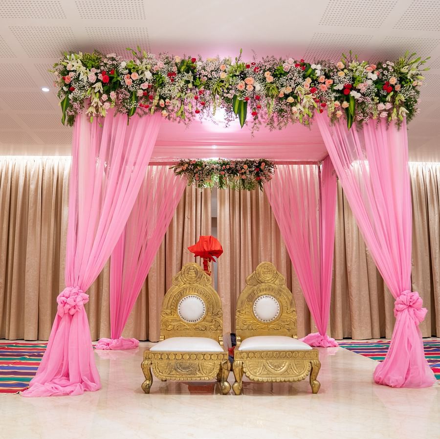 A front view of the wedding mandap decorated with pink drapes, floral arrangements, and a stage with two chairs.