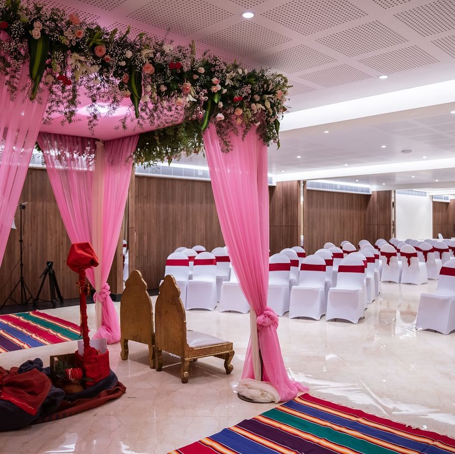  A wedding venue decorated with drapes, floral arrangements, and a stage for the ceremony. Rows of chairs are set up for guests, and there is a carpet leading to the stage.