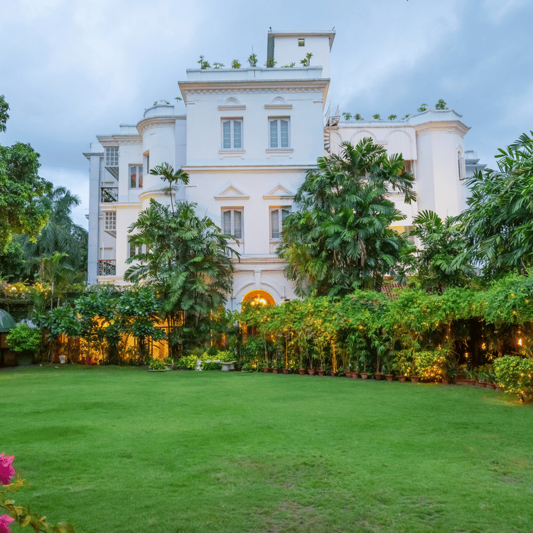 Facade view of Kenilworth Hotel, a city centre hotel in Kolkata, with a lawn in front of it, trees having fairy lights, many trees surrounding the lawn area as seen in late evening.