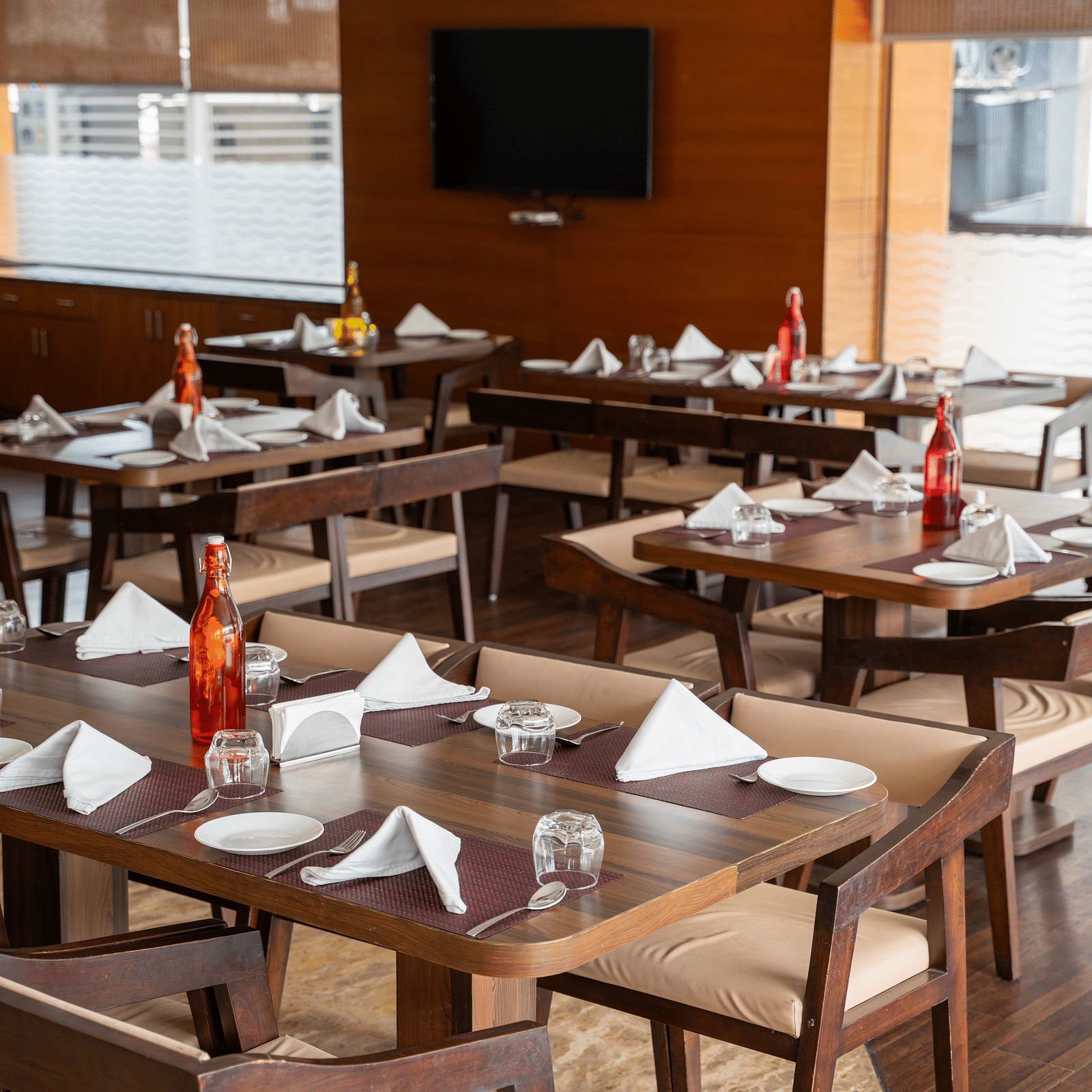 An elegant indoor restaurant setup with polished wooden tables, folded napkins, and large windows providing natural light - Hotel Udai Median, Udaipur.