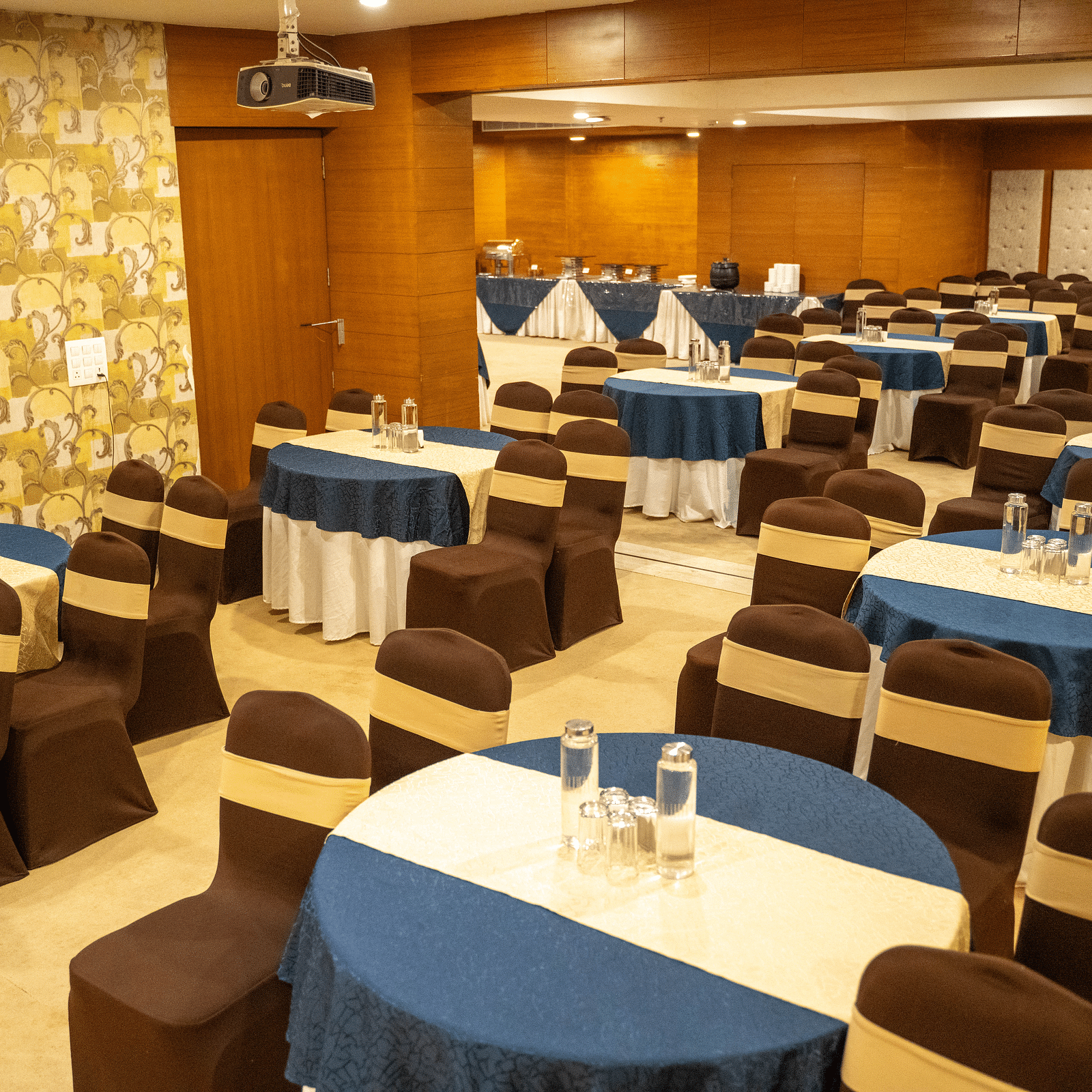 A well-lit event space with round tables, a wooden backdrop, and warm decor - Hotel Udai Median, Udaipur.