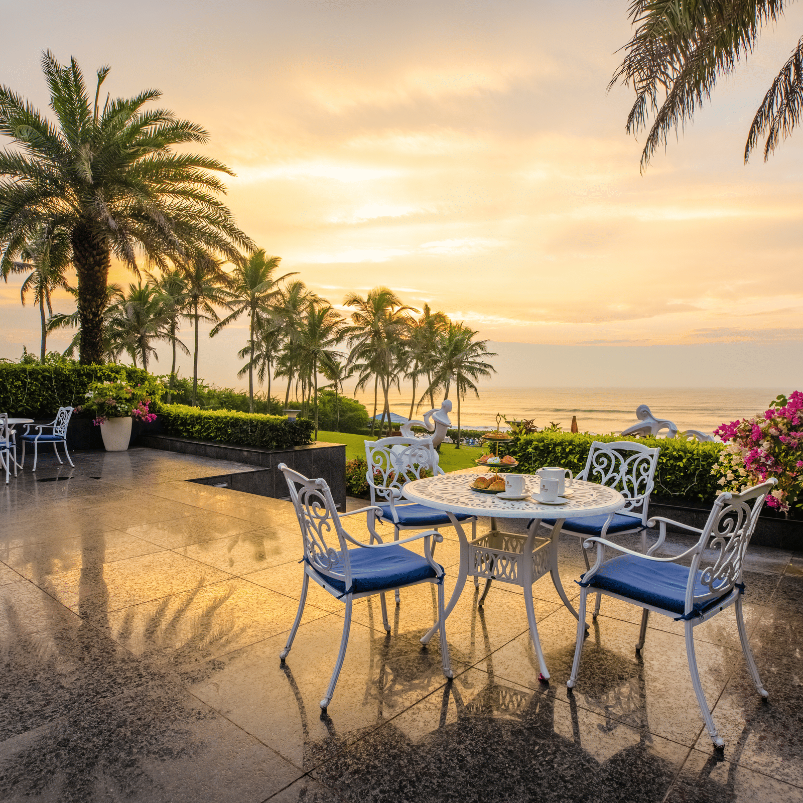 Open tiled terrace with white tables and chairs, clipped hedges and palm trees overlooking the sea at dawn at MAYFAIR Palm Beach Resort, Gopalpur-on-Sea.