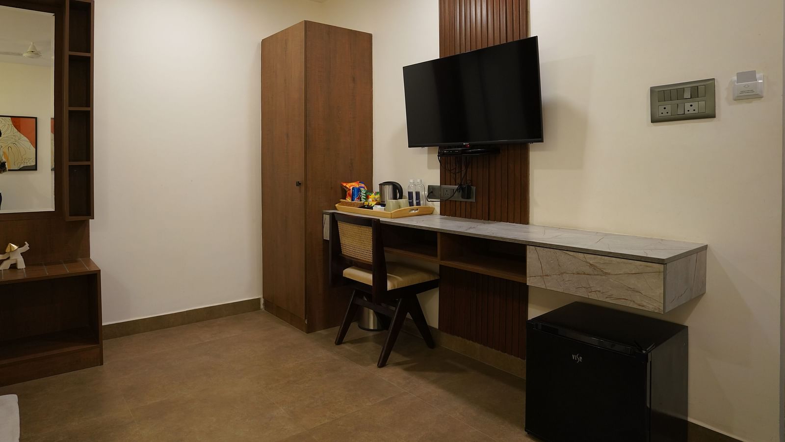 A room with a minimalist design, featuring a wall-mounted TV, a desk with a mirror, and ample lighting at Hotel Kyzen Smartstays – Smart, Reliable, Kyzen