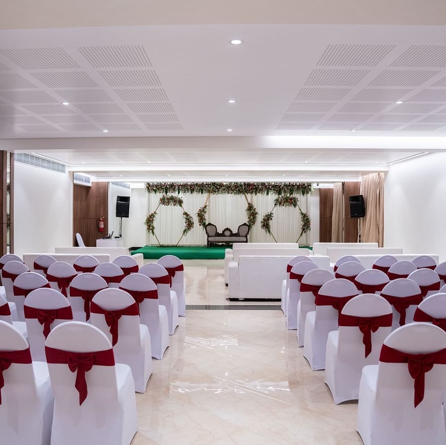 A banquet hall with rows of chairs and a stage, decorated with curtains and floral arrangements.
