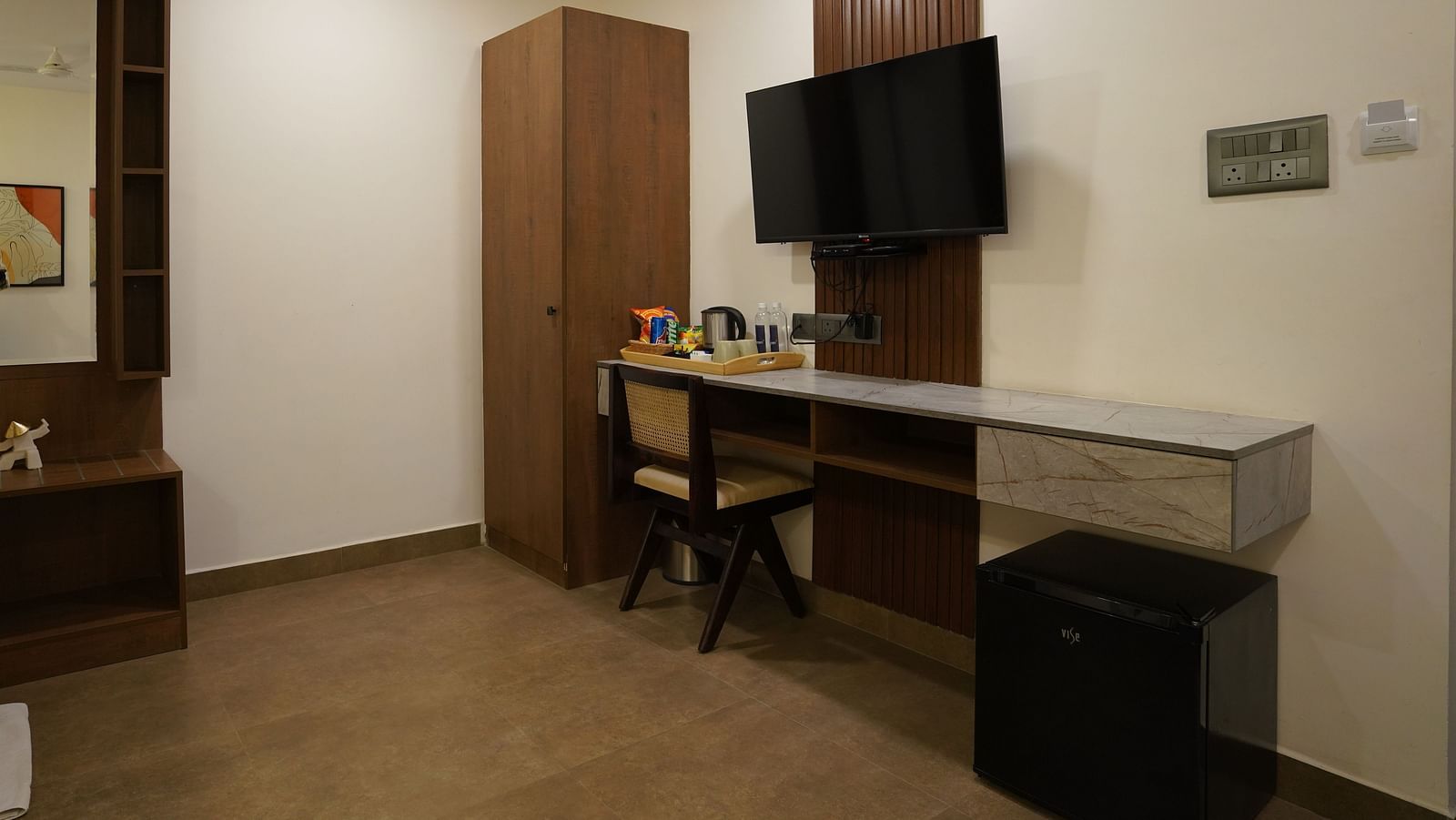 A room with a minimalist design, featuring a wall-mounted TV, a desk with a mirror, and ample lighting at Hotel Kyzen Smartstays – Smart, Reliable, Kyzen
