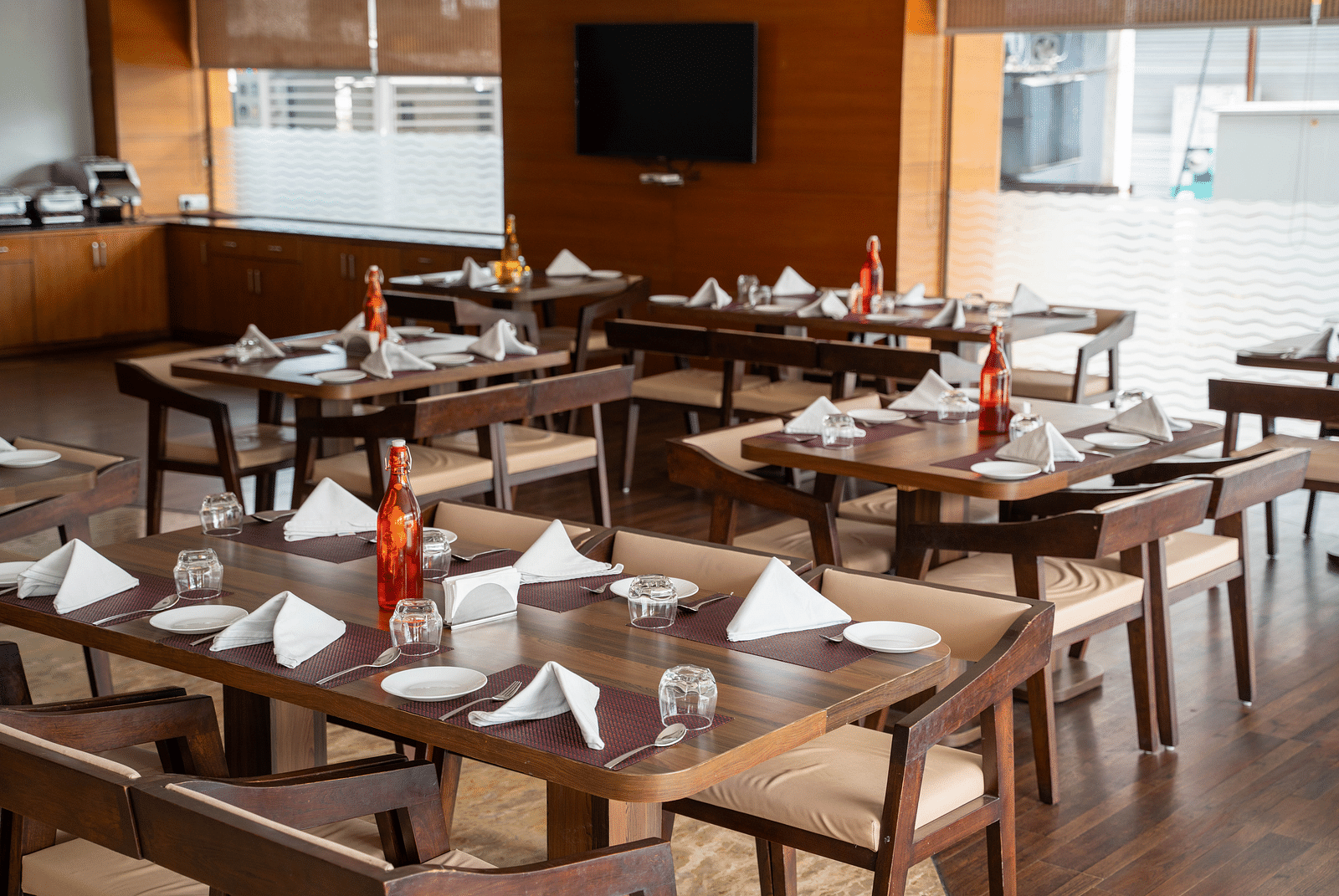 An elegant indoor restaurant setup with polished wooden tables, folded napkins, and large windows providing natural light - Hotel Udai Median, Udaipur.