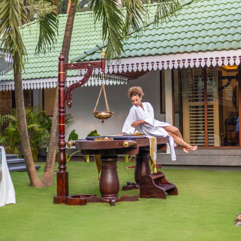 A woman in bathrobe getting on a spa bed which is placed in a lawn of Heritage Village Resort and Spa, Goa. The lawn also has other spa products. 