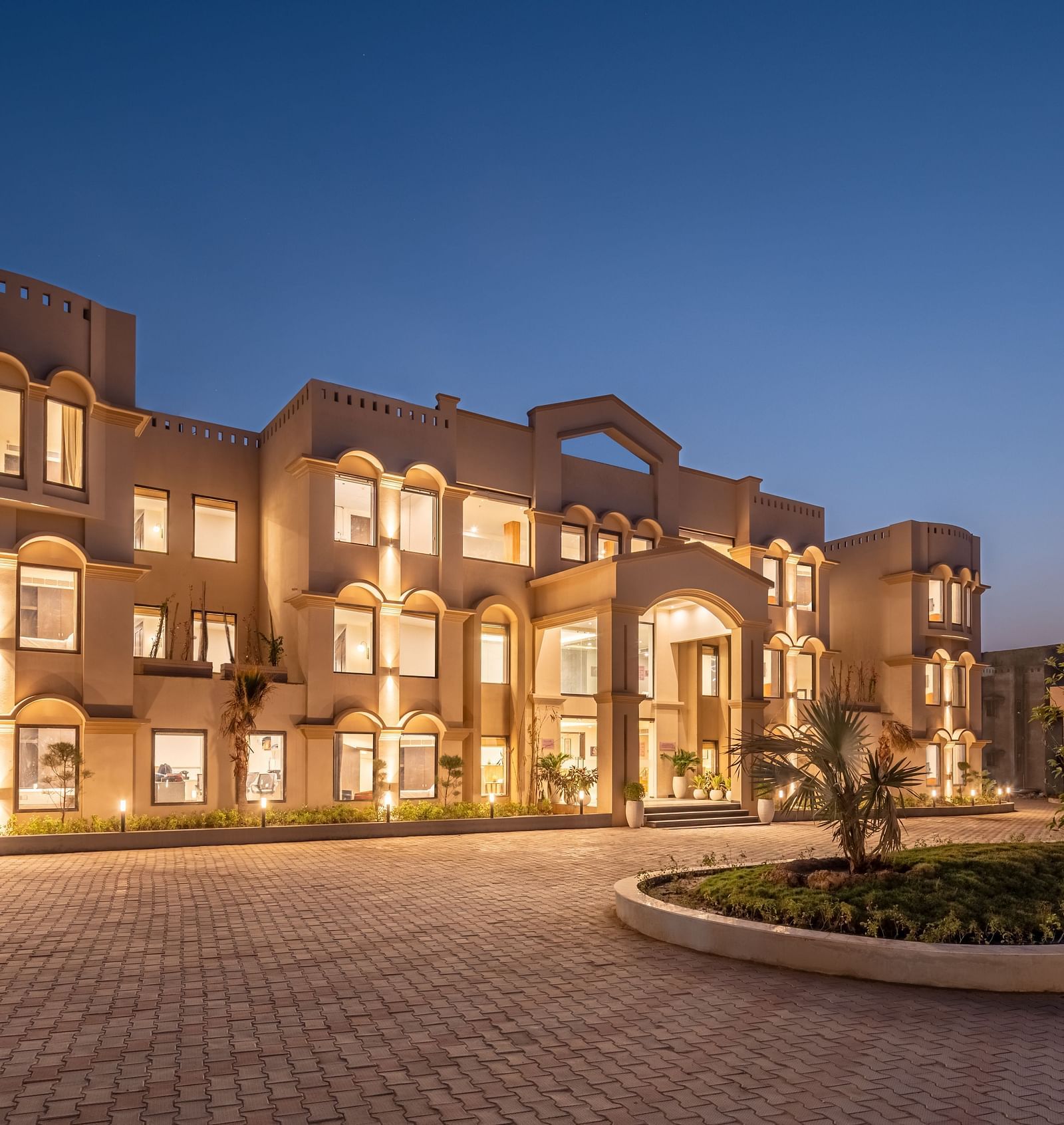 An image of a well lit facade with a huge concrete lawn and a few trees - Brij Eternity, Vrindavan