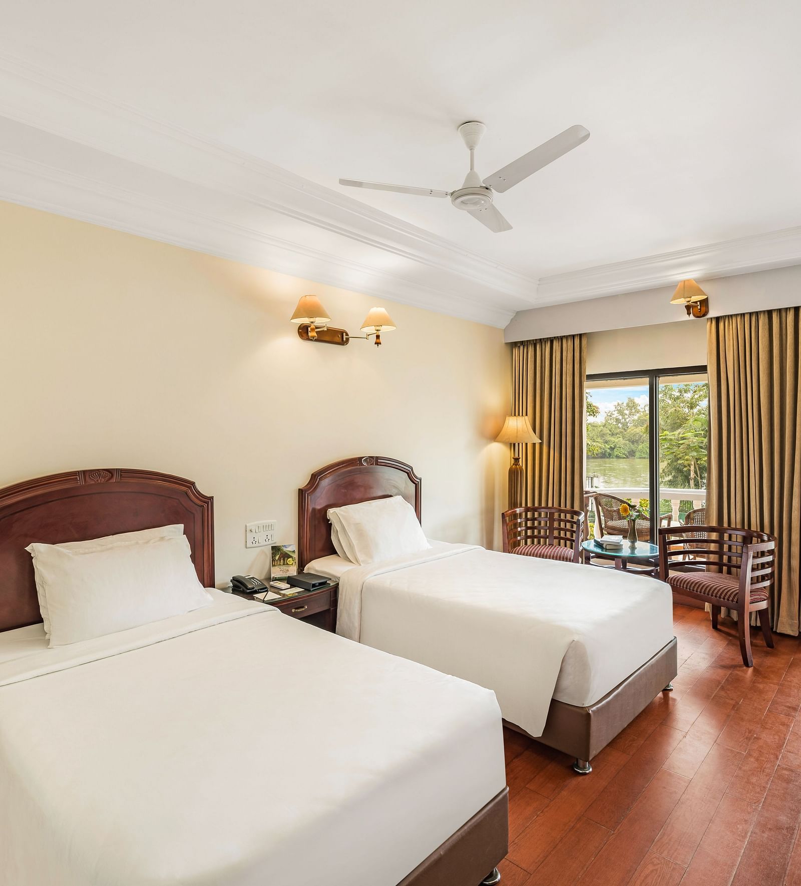 An image of a River View Room with balcony featuring two single beds, bed side table, lamps, ceiling fan, and an expansive glass door leading to the balcony - GReaT Trails River View Resort Thanjavur By GRT Hotels