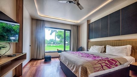 Deluxe Room at Crescent Resort, Sehore featuring a plush bed in front of a TV mounted on the wall and a sitting space next to the large window with the view of the nature outside