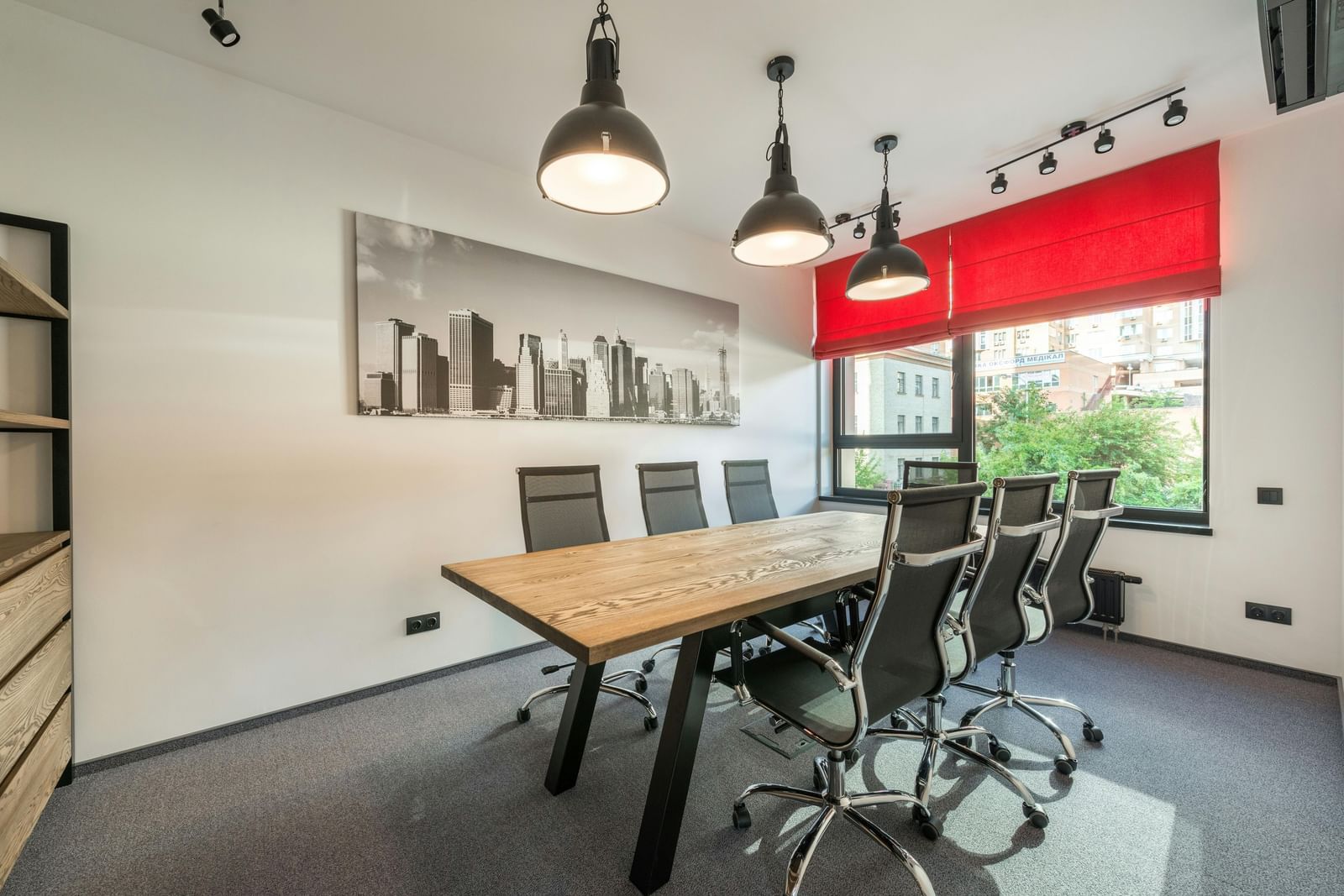 A modern meeting room with a long wooden table, black office chairs, a large photograph, and pendant lights.