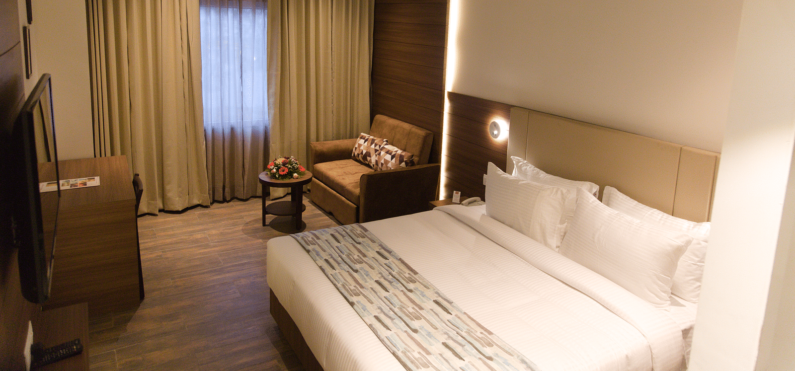 A comfortable hotel room at Lords Inn Udaipur features a double bed with white linens, a cozy armchair by the window, and modern furnishings.