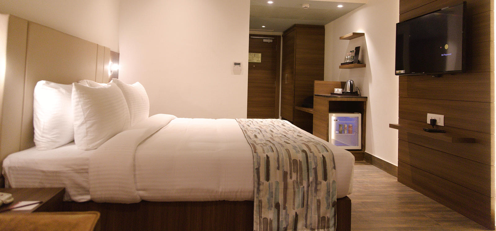 A comfortable hotel room at Lords Inn Udaipur is furnished with a double bed, a mounted television, and a small refrigerator unit.