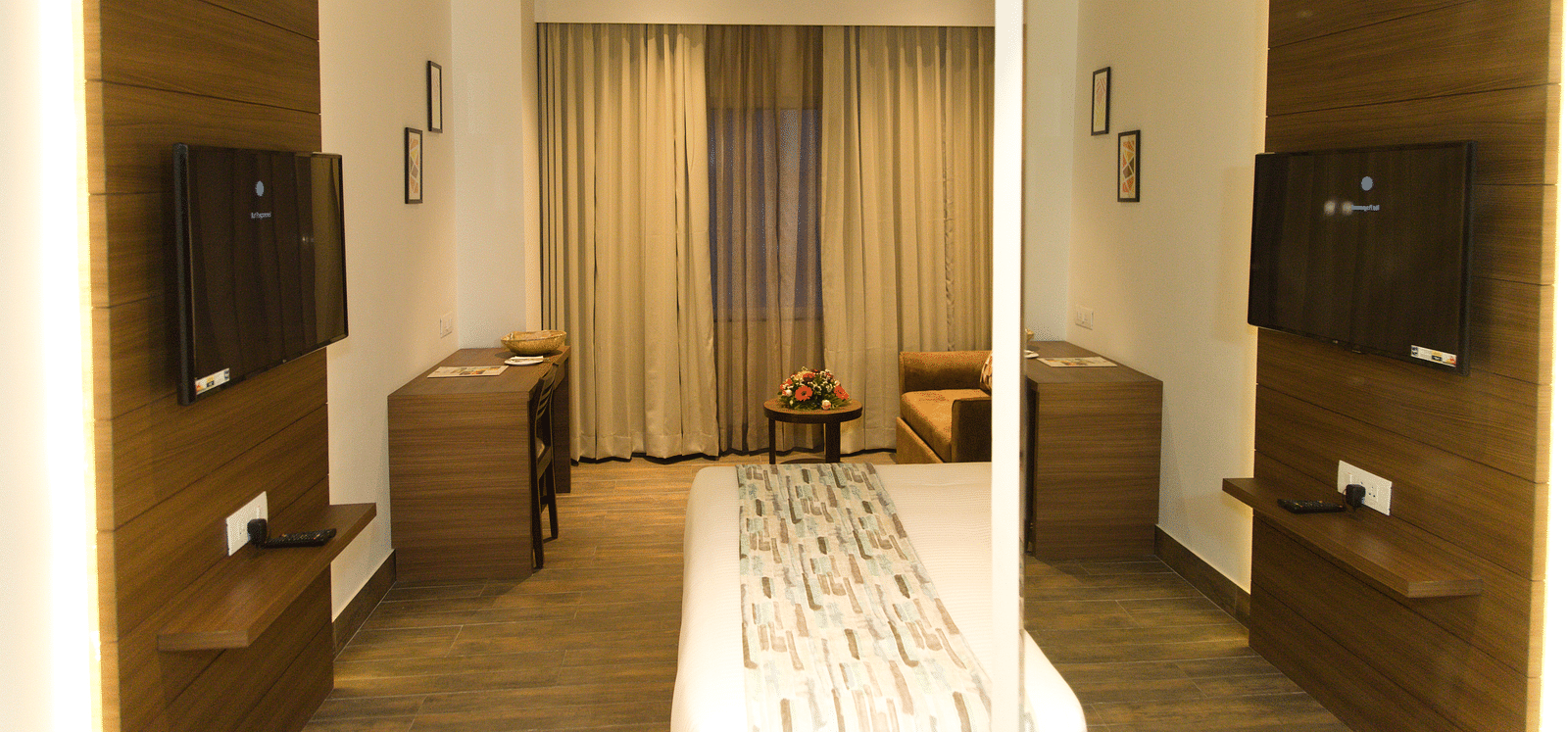 A well-lit hotel room at Lords Inn Udaipur features a bed with a patterned runner, a seating area, and mounted televisions on either side of a partial divider.