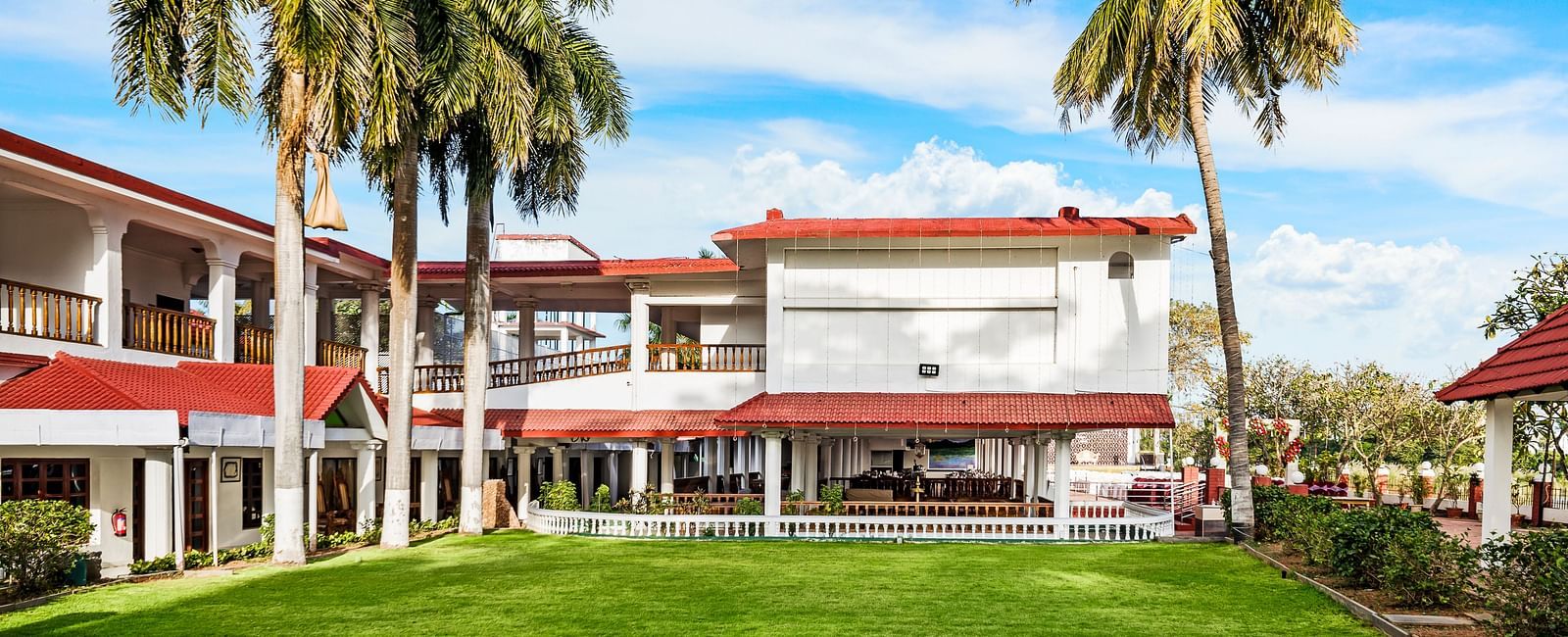 A facade image of the exterior view showcasing a green lawn, tall palm trees with the hotel infrastructure in the background - GReaT Trails River View Resort Thanjavur By GRT Hotels