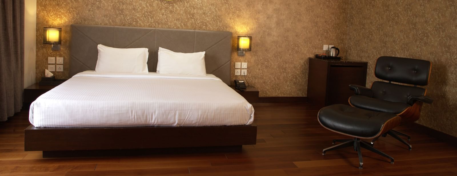 A double bedroom with a lounge chair, a minibar counter, and two bedside lamps at duplicate of a wall-mounted blow dryer and a small mirror at The Hamuse Hotel, Kodaikanal