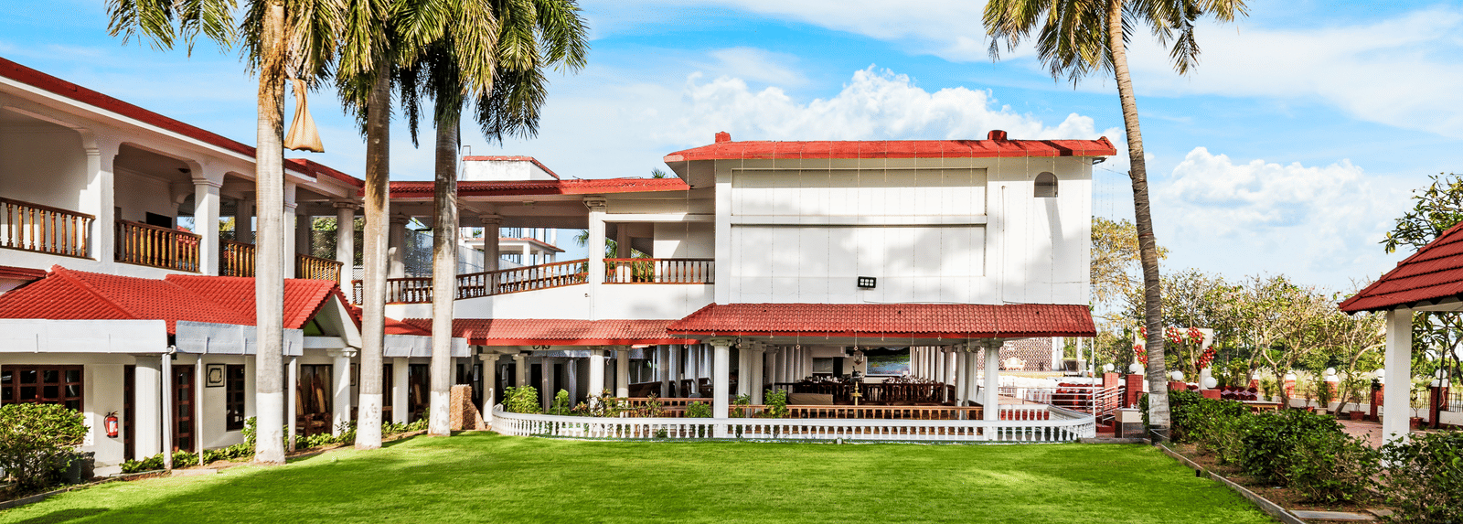 A facade image of the exterior view showcasing a green lawn, tall palm trees with the hotel infrastructure in the background - GReaT Trails River View Resort Thanjavur By GRT Hotels