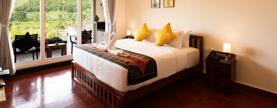 A bed with cushions and a runner sits on a polished floor inside the Bison room at Ibex Resort, Kotagiri. Glass doors with curtains lead to a balcony area. Frames hang on the wall above the bed structure.