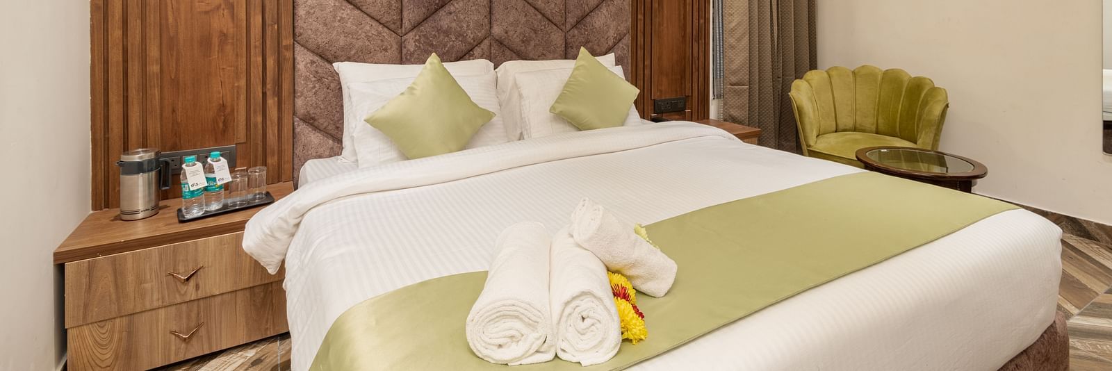 Image of Deluxe Room at DLS Grand Luxotica, Dehradun featuring a plush bed with rolled bath towels placed on the bed runner and a sitting space with a teapoy and couch next to it