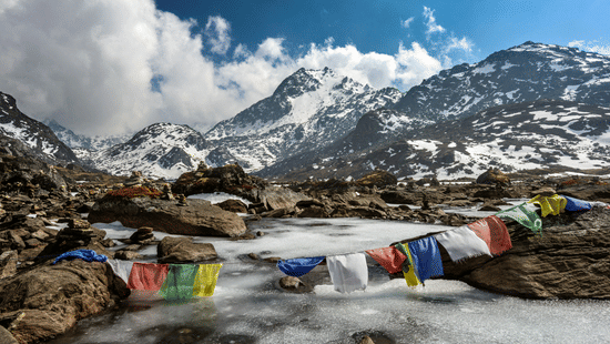 A snowy mountain landscape with a partially frozen stream and colorful prayer flags in the foreground.