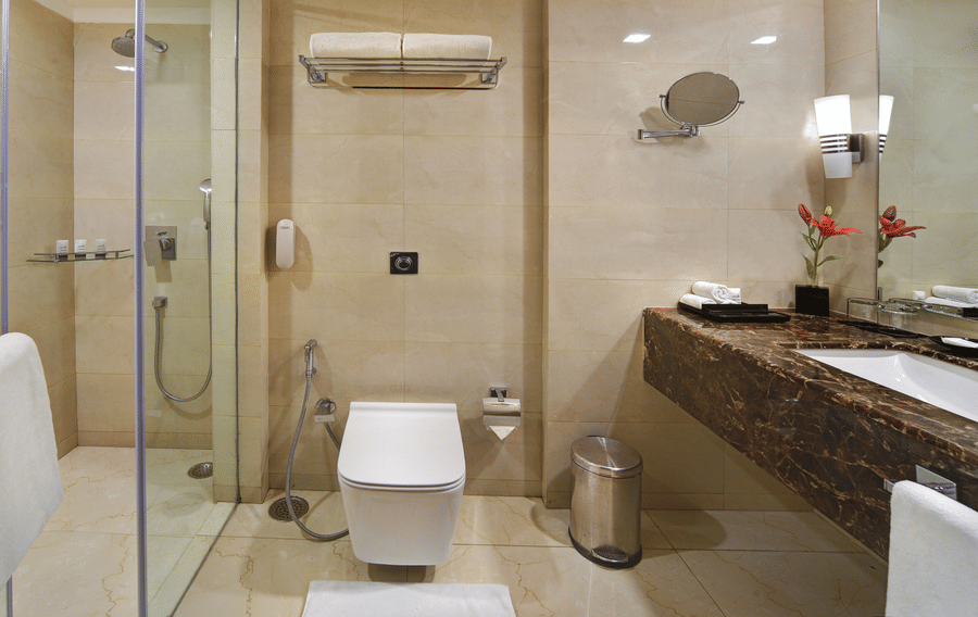 Bathroom at The Bristol Hotel, Gurgaon with beige marble tiles, a glass-enclosed shower, a wall-mounted toilet, and a dark marble vanity counter with a mirror.