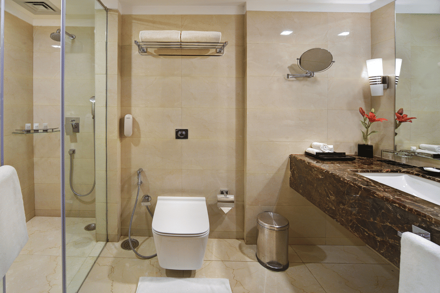 Bathroom at The Bristol Hotel, Gurgaon with beige marble tiles, a glass-enclosed shower, a wall-mounted toilet, and a dark marble vanity counter with a mirror.