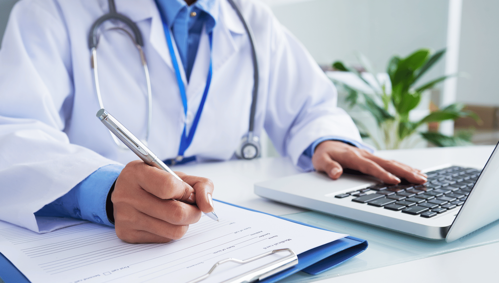 Doctor in a white coat writing on a clipboard while using a laptop, seated at a desk.
