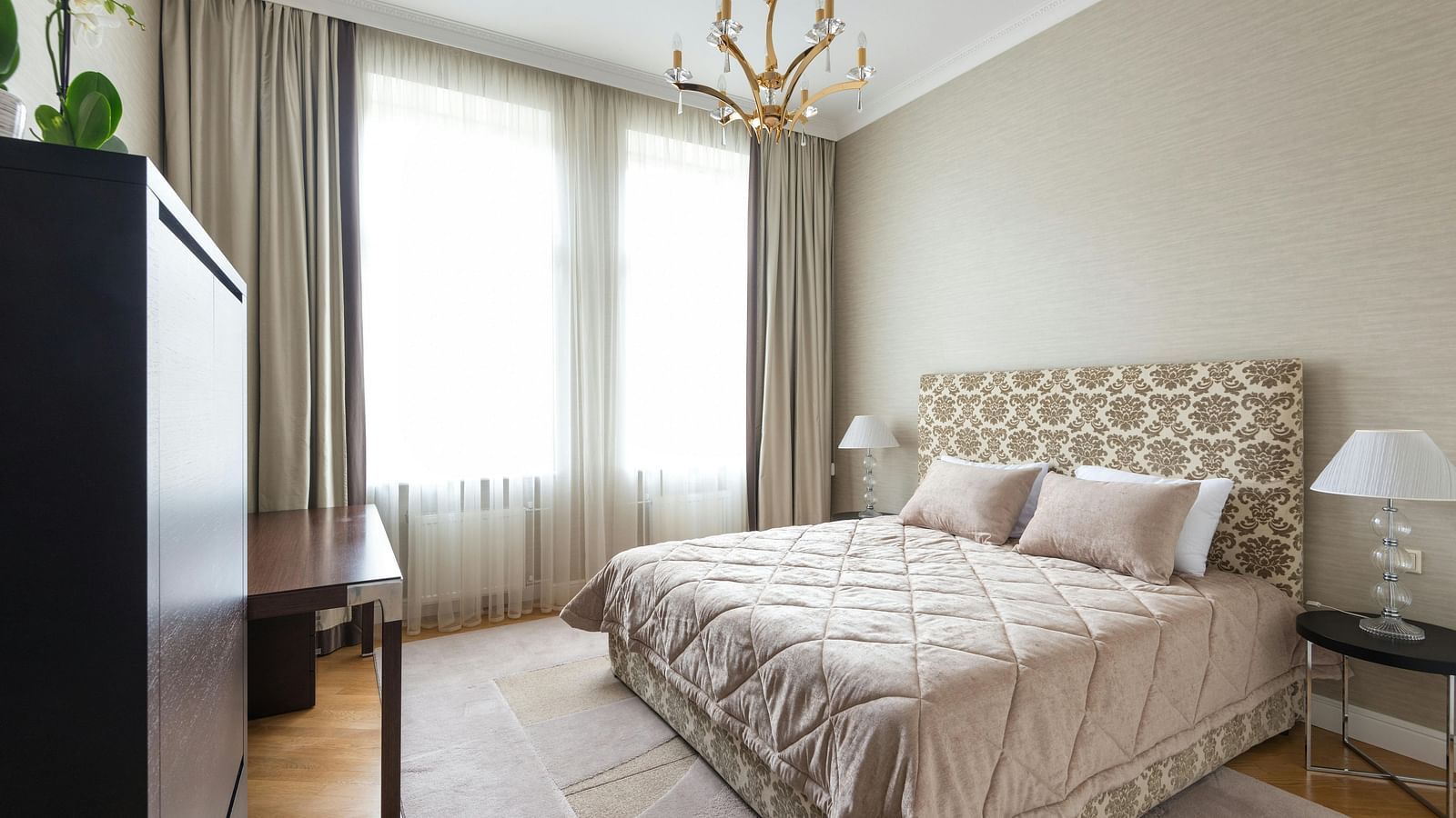 A bedroom features a patterned bedspread, neutral walls, and a chandelier.