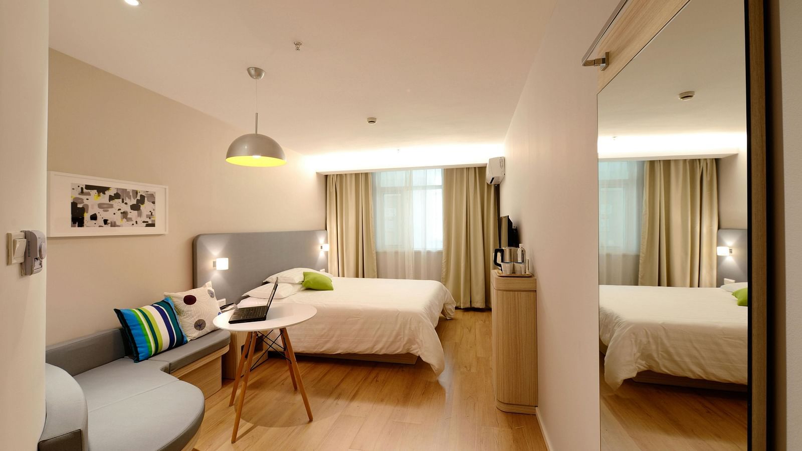 A bright hotel suite includes a seating area and a bed, reflected in a large mirror.