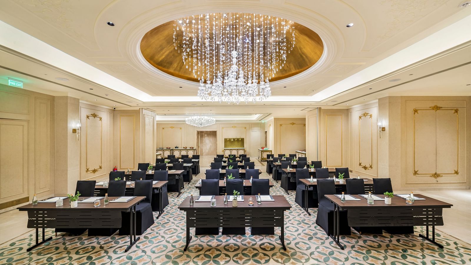 A grand ballroom configured for a large seminar or presentation at Grand Chennai by GRT Hotels showcasing numerous black tables and chairs neatly arranged in rows. A magnificent, large chandelier dominates the high ceiling, enhancing the luxurious feel.