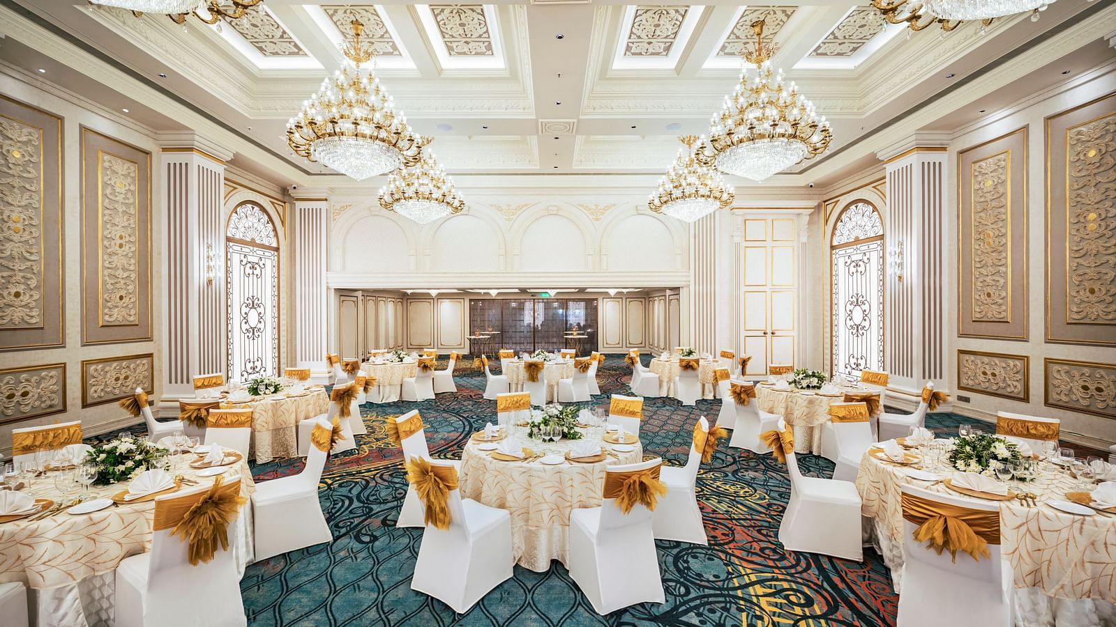 A grand hall prepared for a reception at Grand Chennai by GRT Hotels  showcasing elegant round tables with golden accents and white chairs, all beneath a ceiling adorned with multiple sparkling chandeliers.
