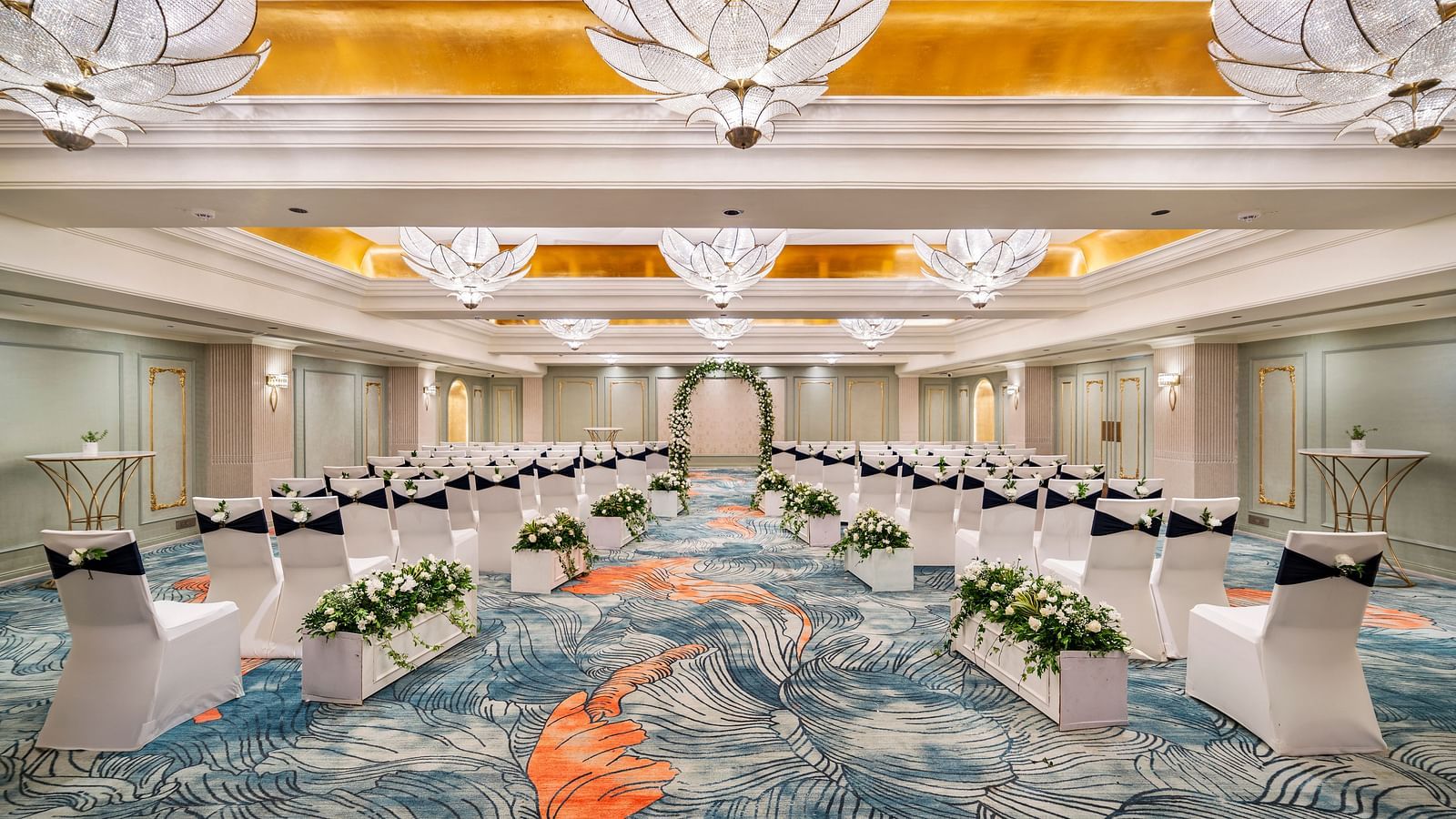 An elegant ballroom at Grand Chennai by GRT Hotels is set for a formal gathering, showcasing meticulously aligned white chairs, tasteful floral centerpieces, and magnificent, decorative chandeliers that illuminate the entire space.